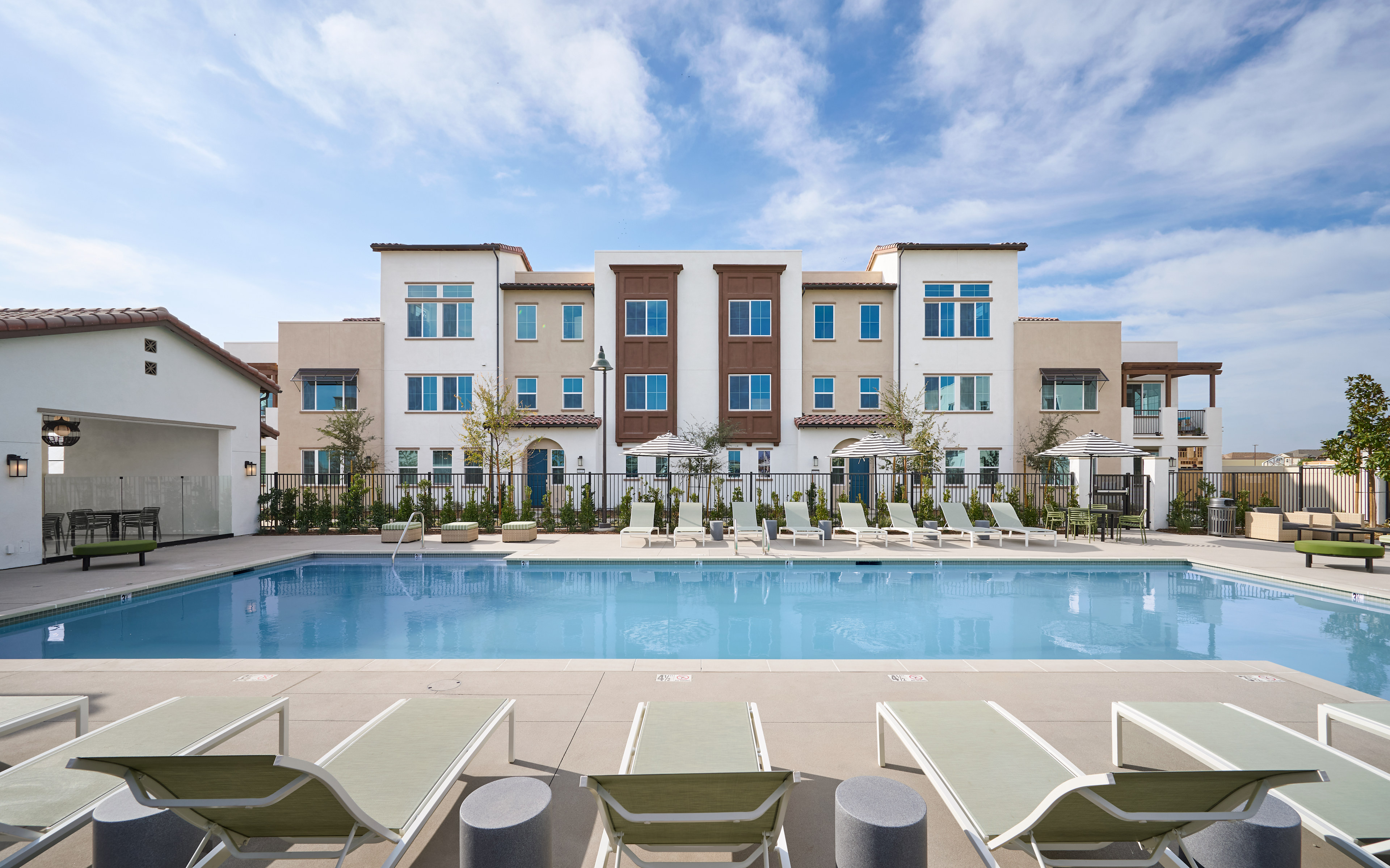 A modern multi-story apartment building with a swimming pool in the foreground, surrounded by lounge chairs and a lush outdoor environment under a bright, cloudy sky.