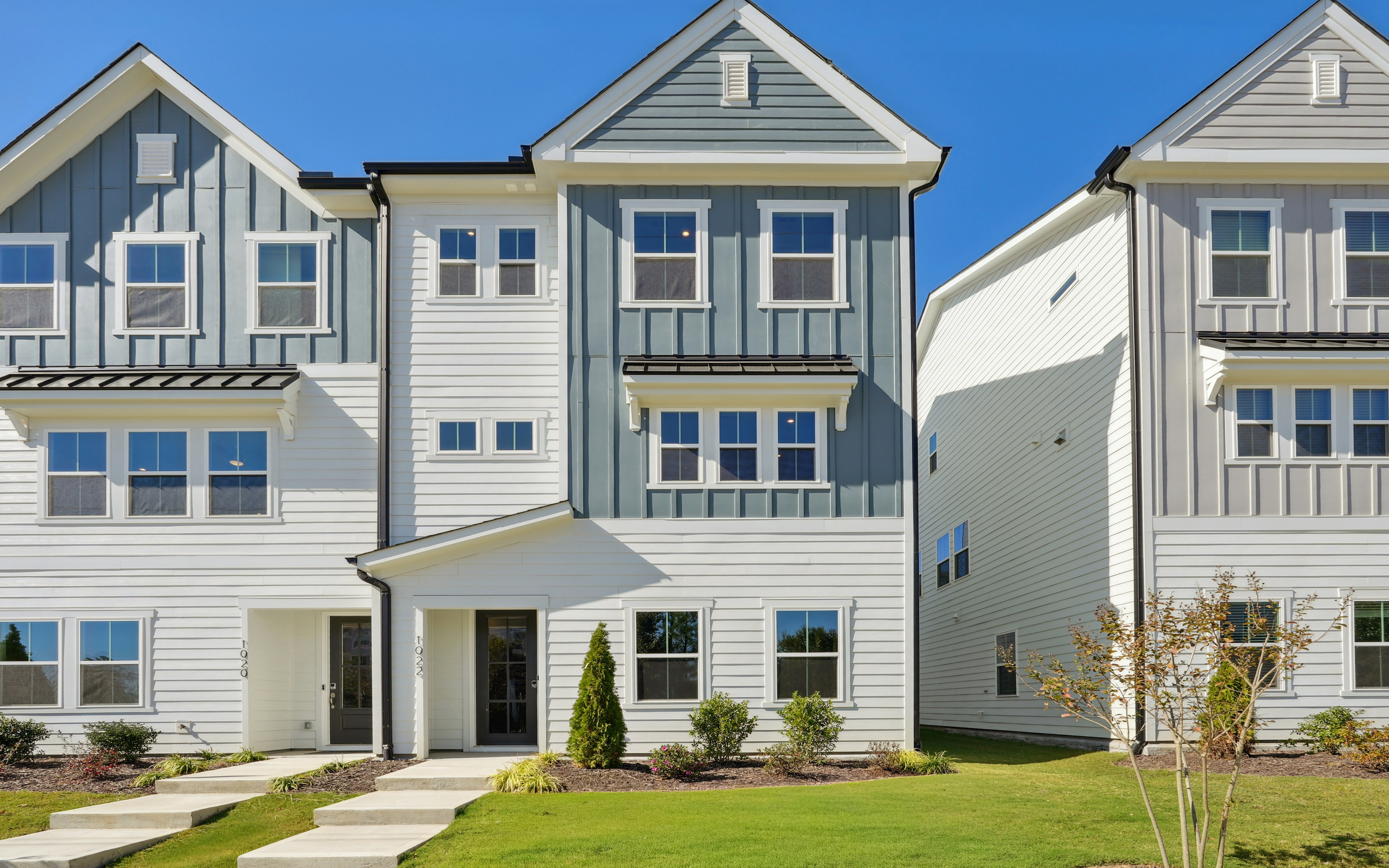 A row of modern, two-story townhouses with gray siding, white trim, and gabled roofs set against a clear blue sky, surrounded by lush greenery in the foreground.