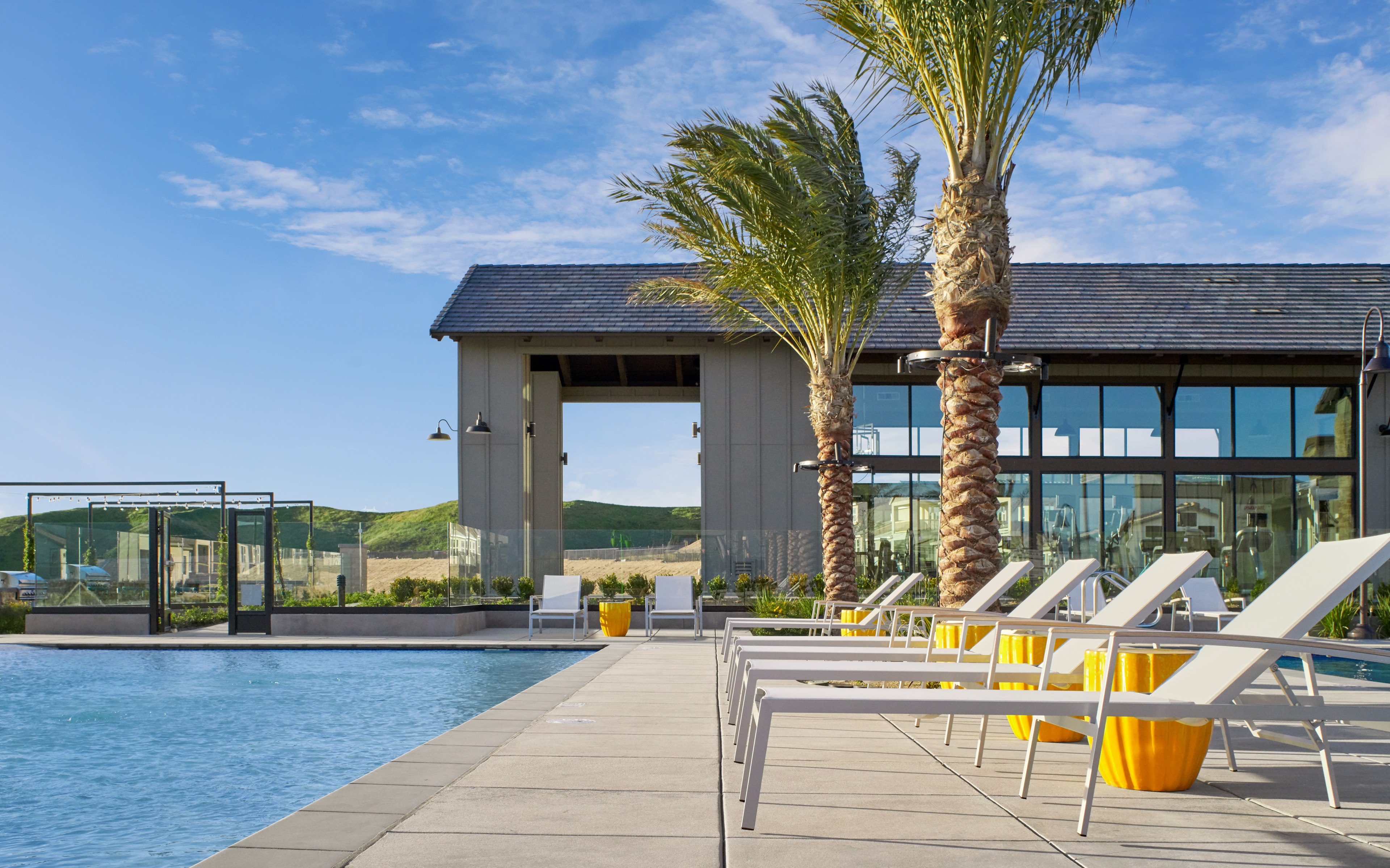 A modern outdoor pool area with palm trees, lounge chairs, and a glass-walled building in the background against a clear blue sky.