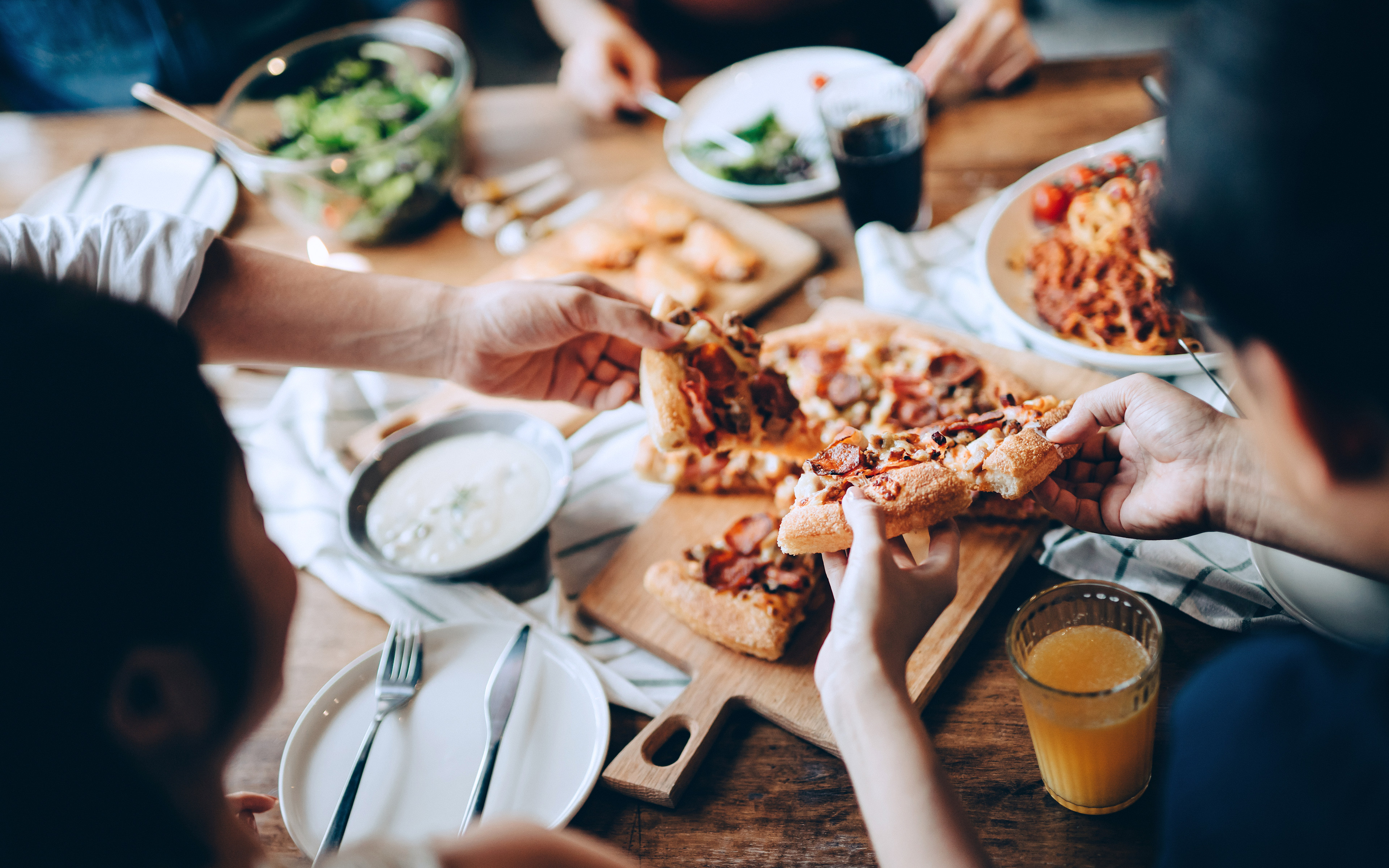 A group of people sharing a meal, with various dishes and drinks spread out on a wooden table in the foreground, while the background appears to be a dimly lit setting.