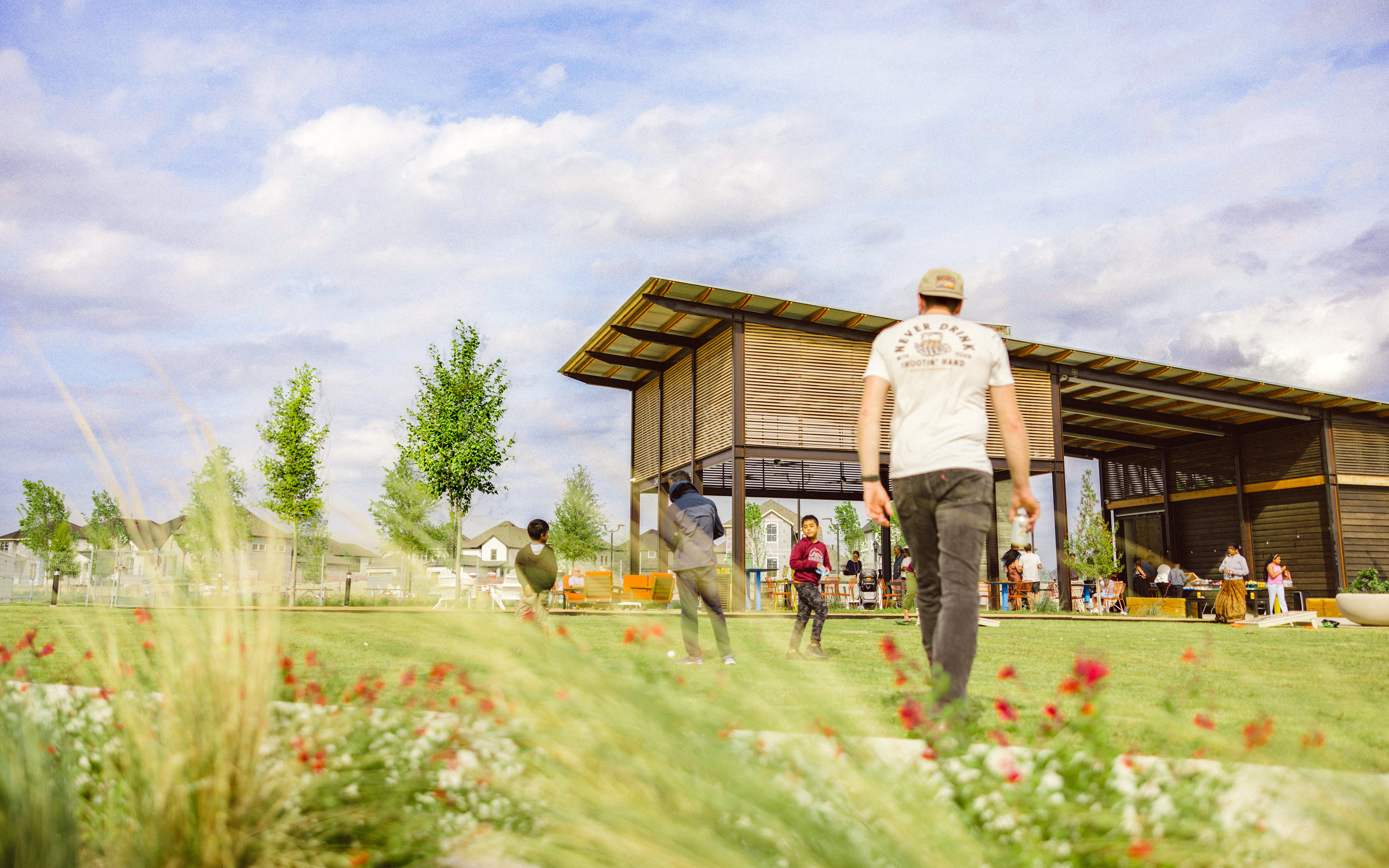 A wooden structure with a covered porch stands in a grassy field, surrounded by blooming red flowers and lush greenery, with a cloudy sky overhead.