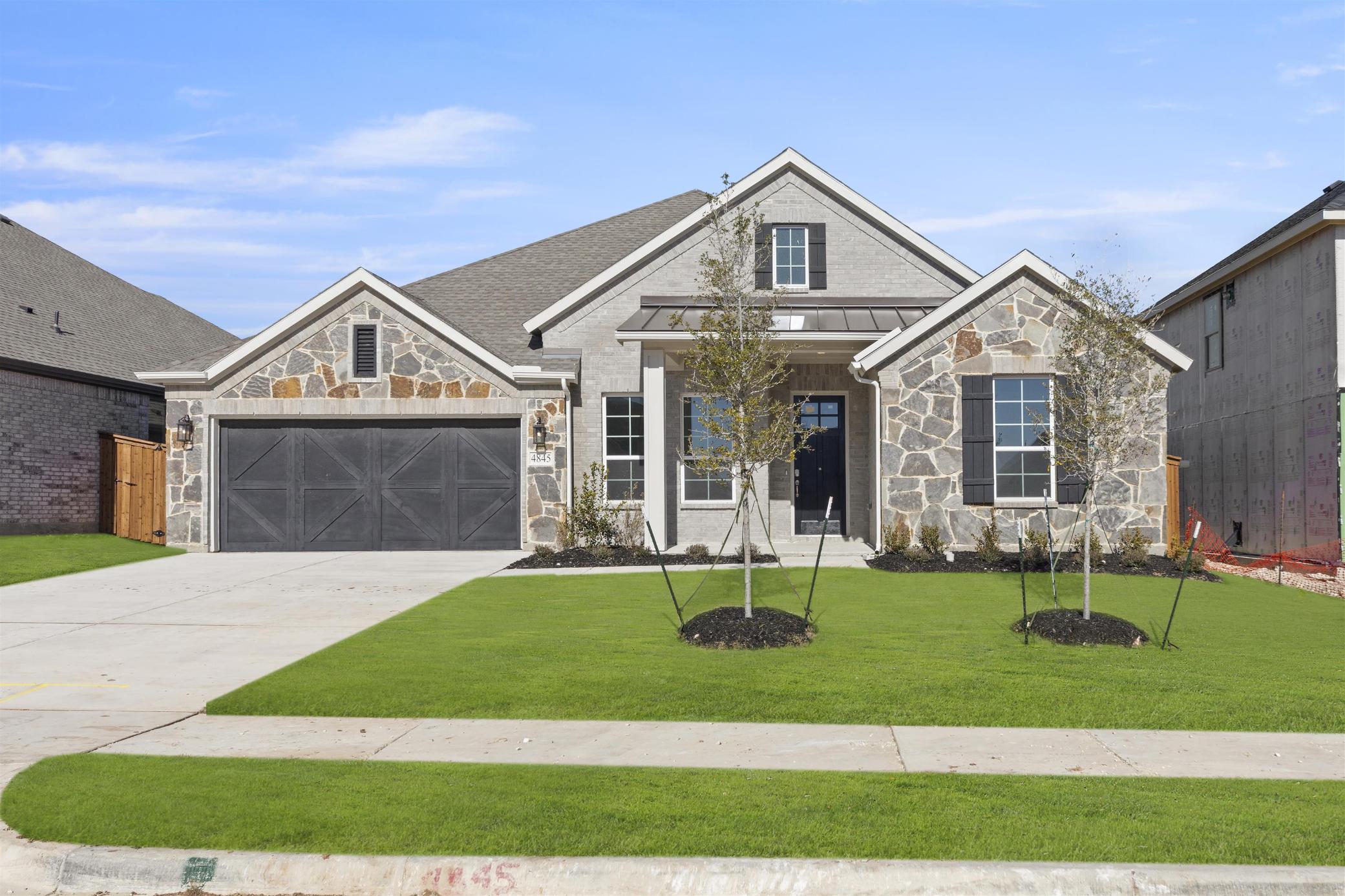 A two-story residential house with a stone exterior, a gabled roof, and a well-manicured lawn in the foreground, set against a clear blue sky with some clouds.