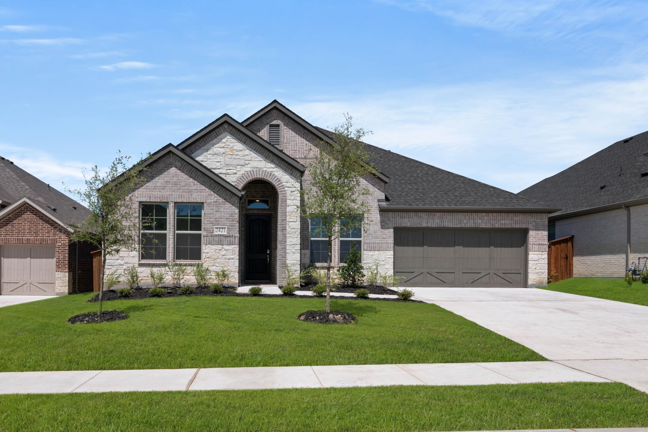 A well-manicured lawn surrounds a two-story brick house with a gabled roof, set against a clear blue sky with scattered clouds.