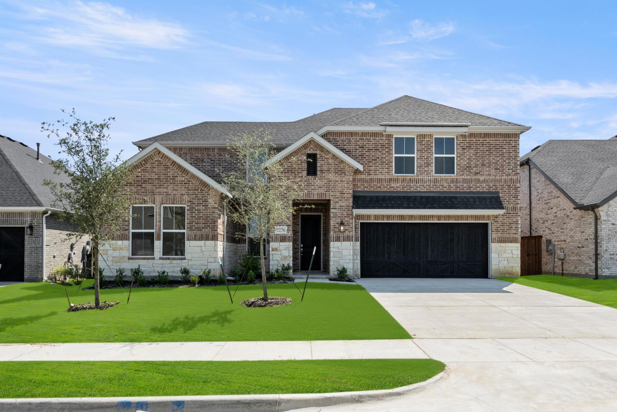 A two-story brick house with a well-manicured lawn and a driveway leading to a garage door in the foreground, set against a clear blue sky with scattered clouds.