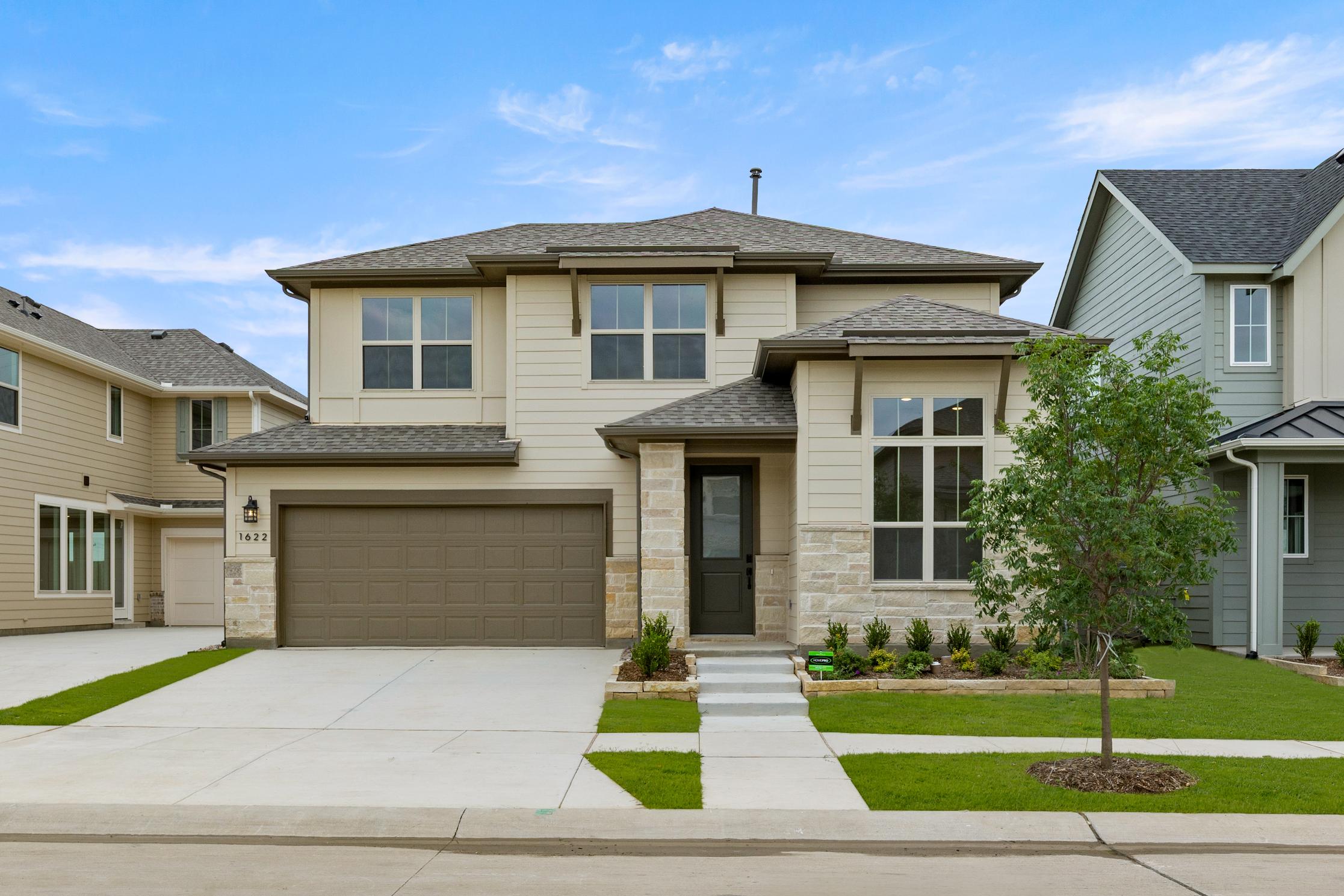 A two-story beige house with a garage, surrounded by a well-manicured lawn and landscaping, stands against a clear blue sky with scattered clouds.