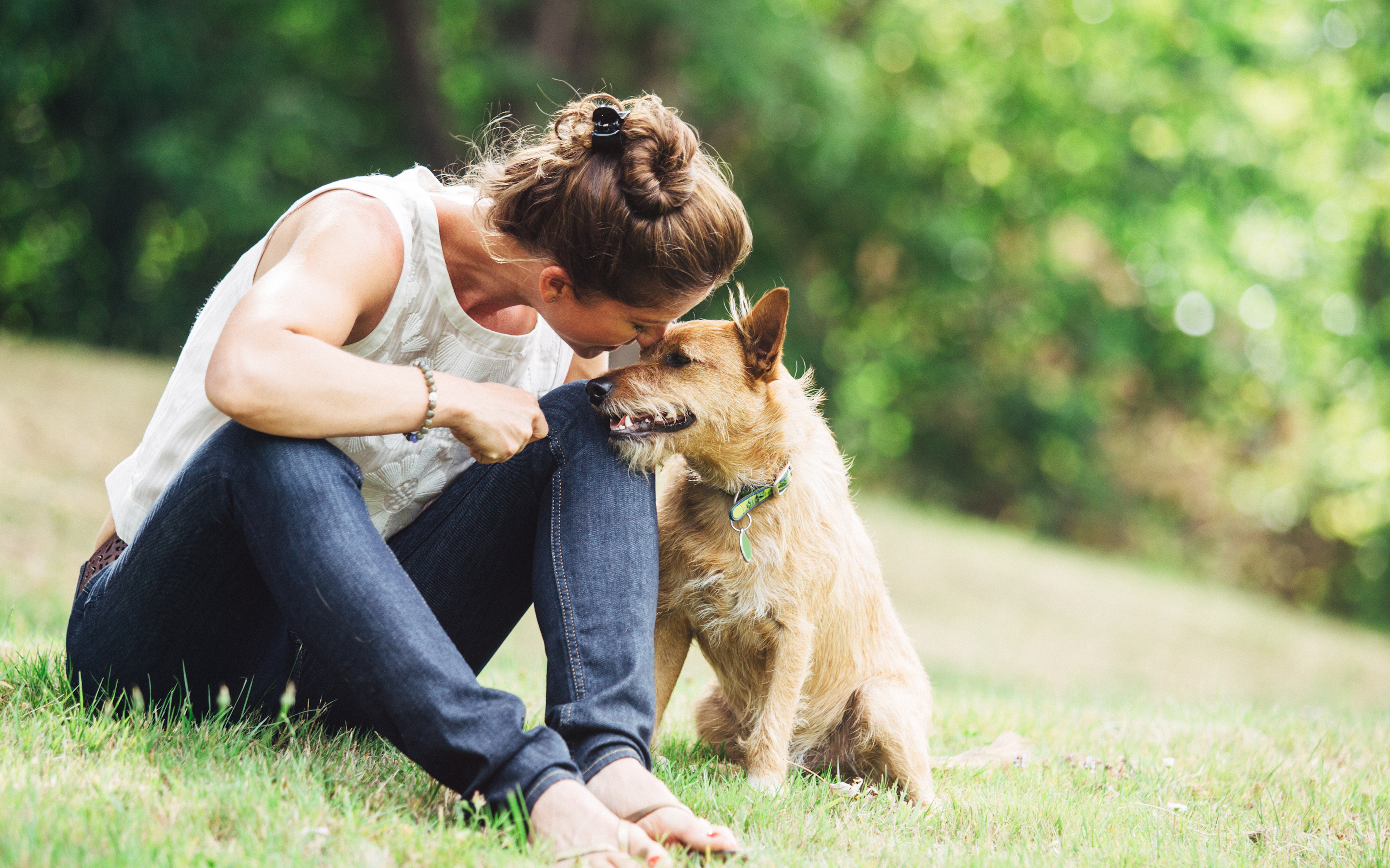 A woman in a white tank top is sitting on the grass, embracing a German shepherd dog in a lush, green outdoor setting.