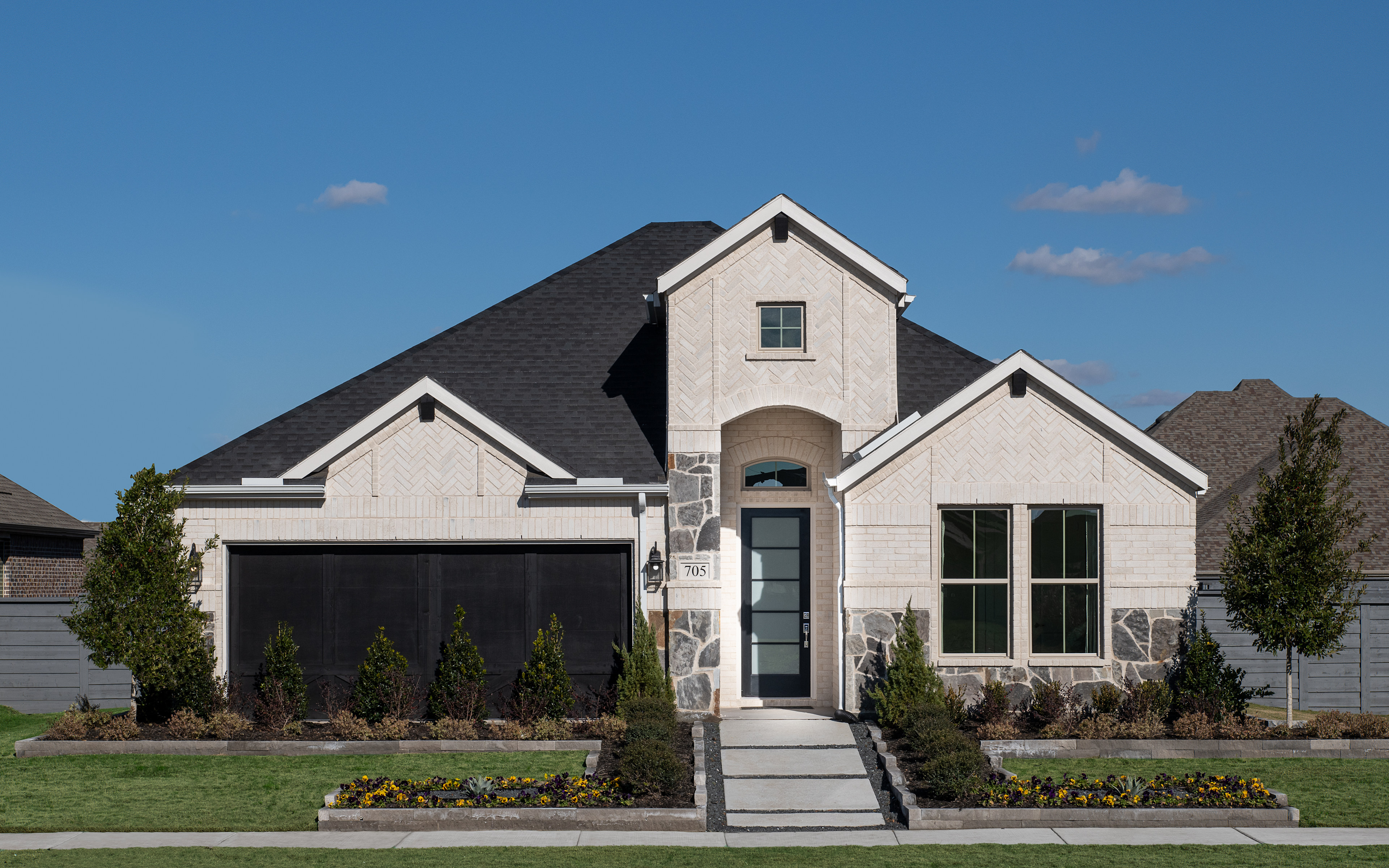 A two-story house with a gabled roof, stone and siding exterior, and a landscaped front yard with shrubs and flowers, set against a clear blue sky with a few clouds.