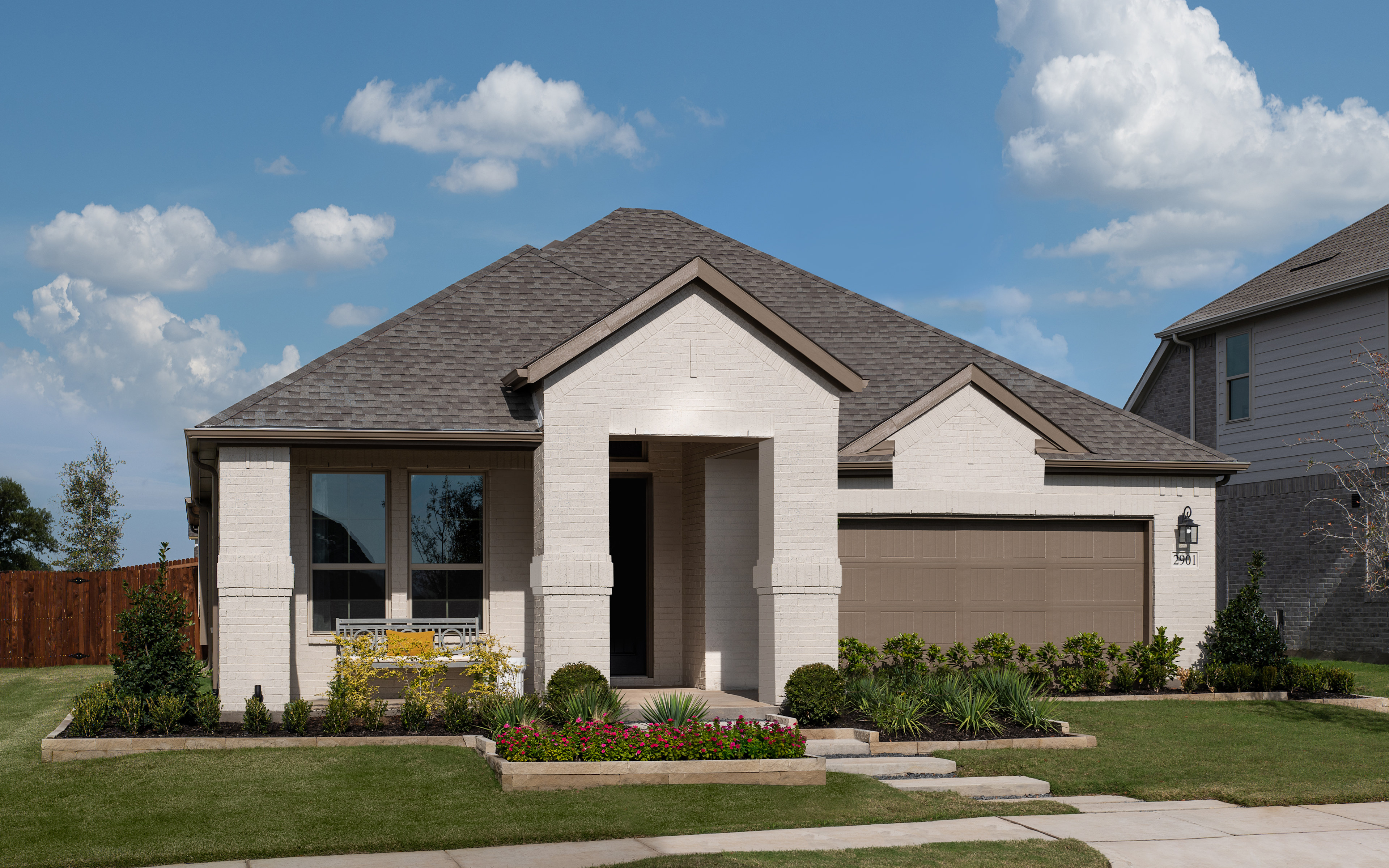 A well-manicured lawn surrounds a two-story house with a gabled roof, stone and stucco exterior, and a covered porch leading to the entrance. The sky is clear with fluffy white clouds, creating a picturesque suburban scene.