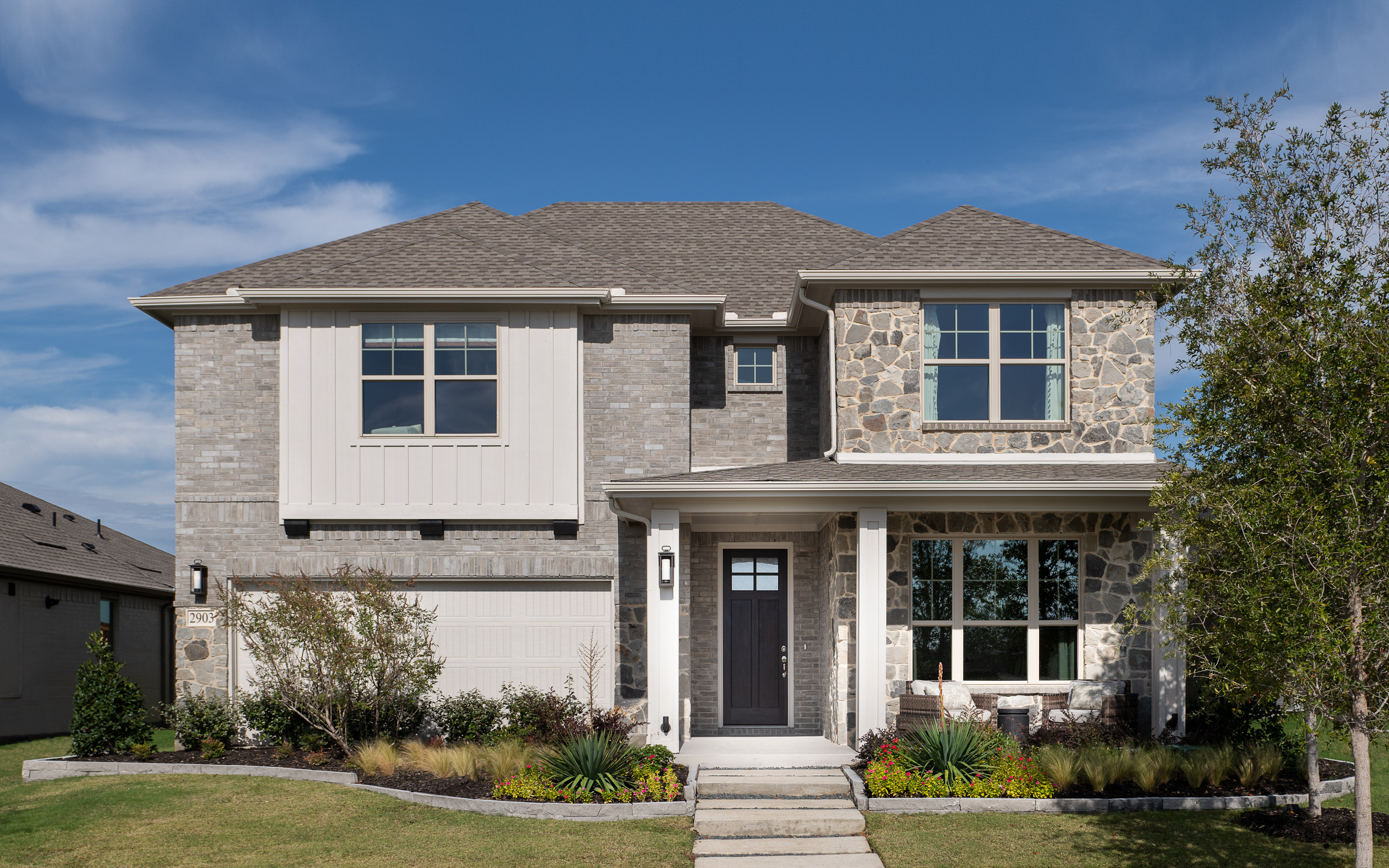 A two-story residential house with a stone and stucco exterior, surrounded by a well-manicured lawn and landscaping, set against a clear blue sky with scattered clouds.