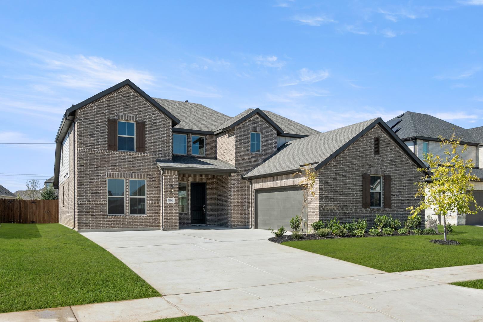 A large, two-story brick house with a well-manicured lawn and a concrete driveway in the foreground, set against a clear blue sky with scattered clouds.