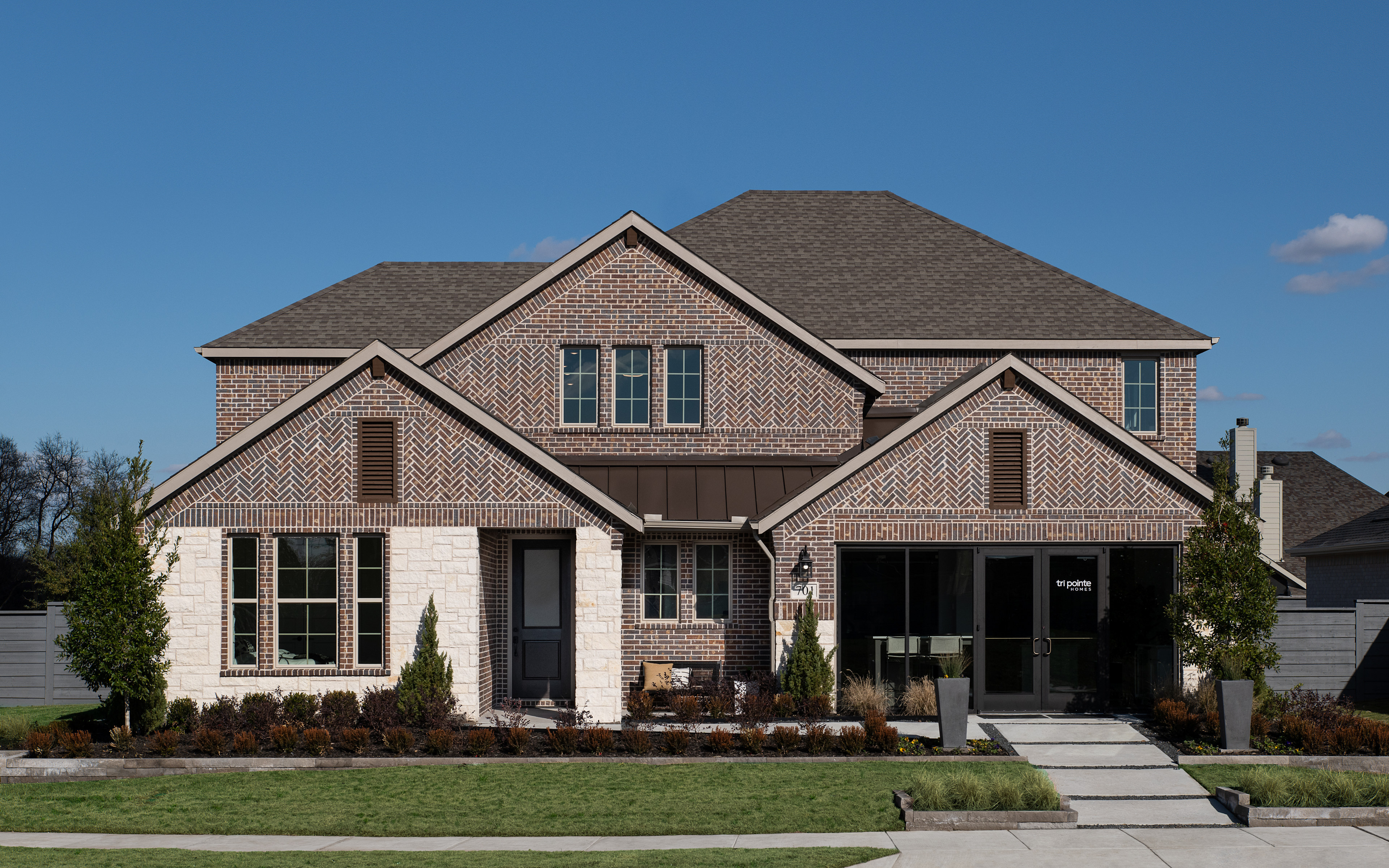 A two-story brick house with a gabled roof, surrounded by landscaped gardens and a paved driveway, set against a clear blue sky.