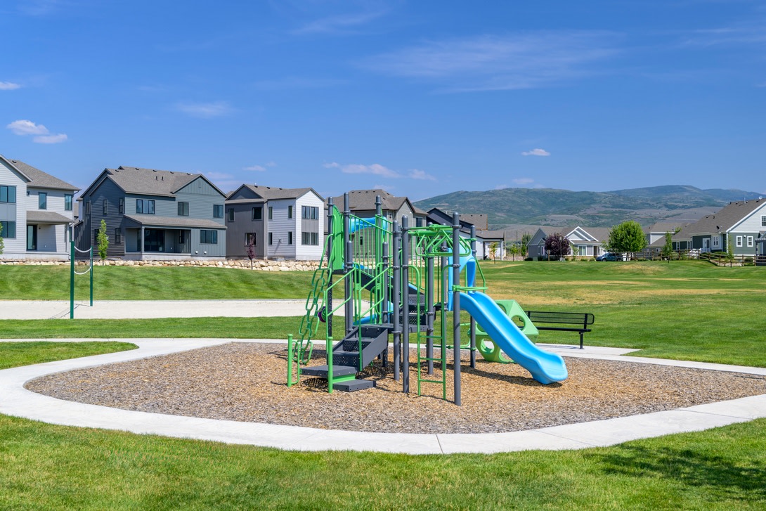 A colorful playground with slides and climbing structures sits in a grassy area, surrounded by a residential neighborhood with houses and mountains in the background.