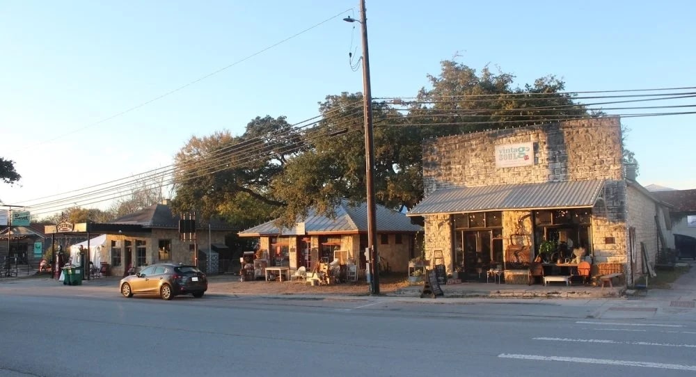 A quaint street scene with a row of small shops and businesses, surrounded by trees and power lines, with a car parked on the street in the foreground.