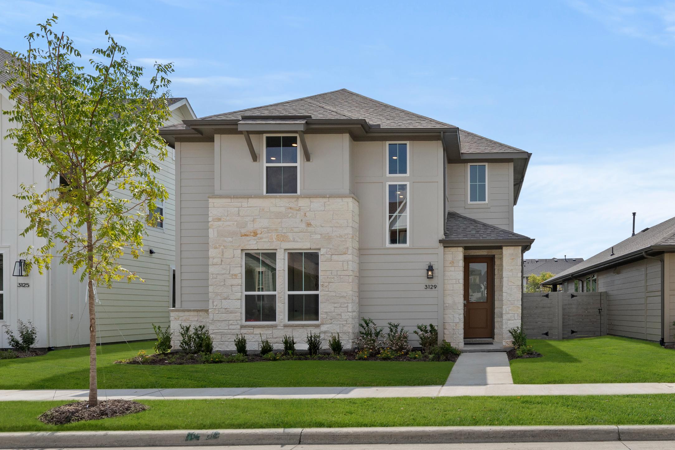 A two-story residential house with a stone exterior, surrounded by a well-manicured lawn and landscaping, set against a clear blue sky.