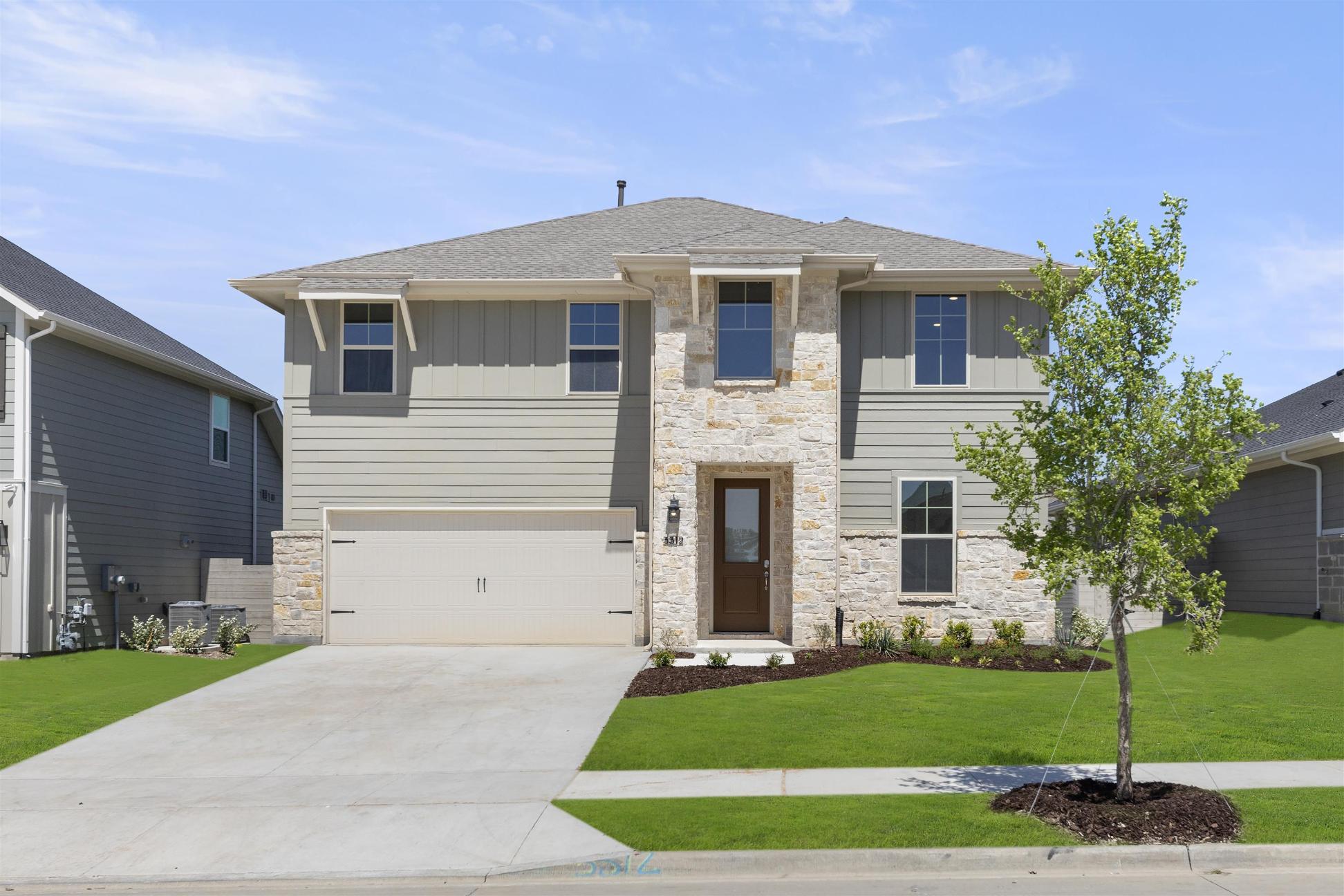 A two-story residential house with a stone exterior, a tiled roof, and a well-manicured lawn in the foreground, set against a clear blue sky with some trees in the background.