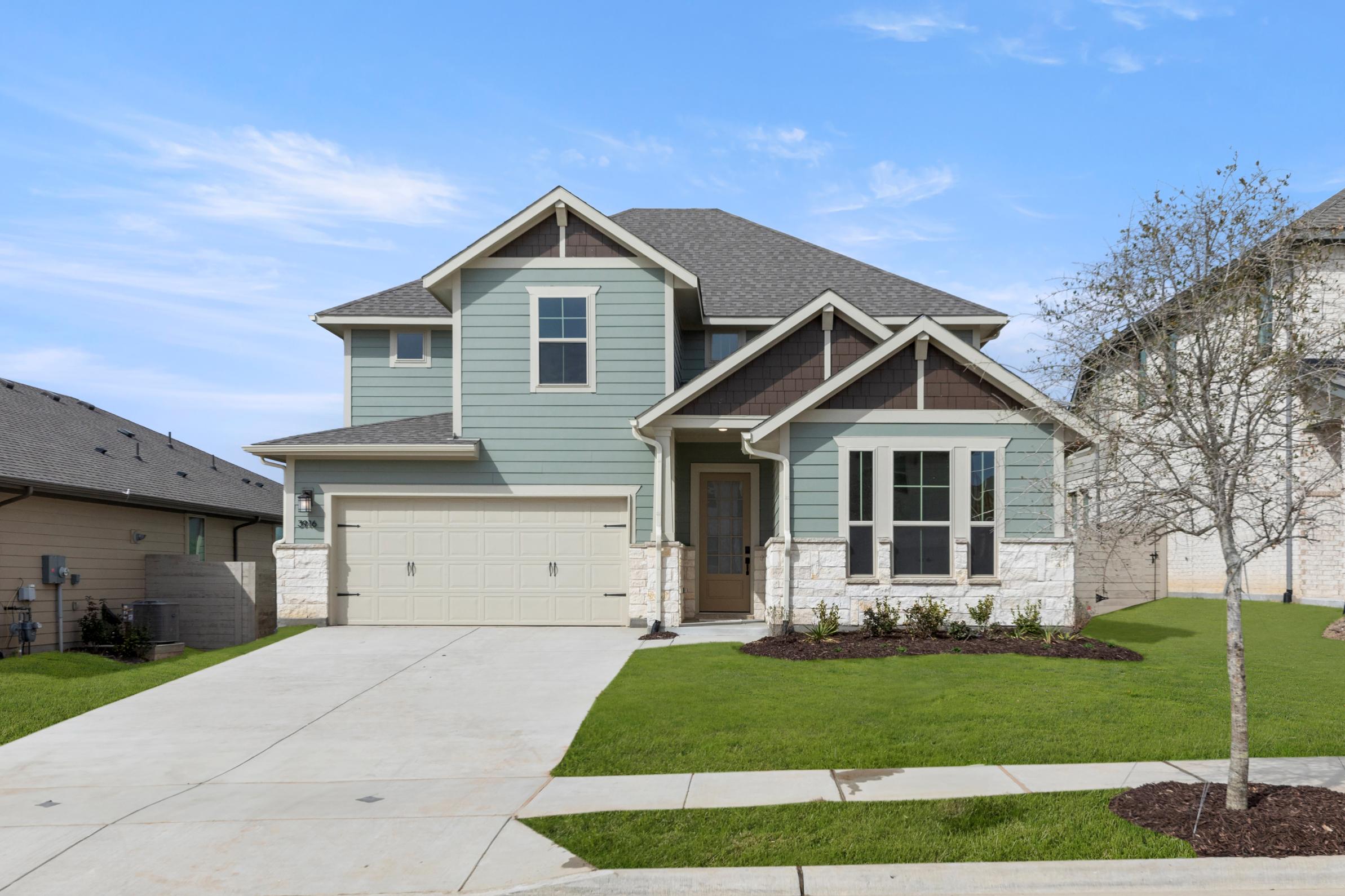 A two-story house with a green exterior, a gabled roof, and a paved driveway in the foreground, set against a clear blue sky with some clouds.