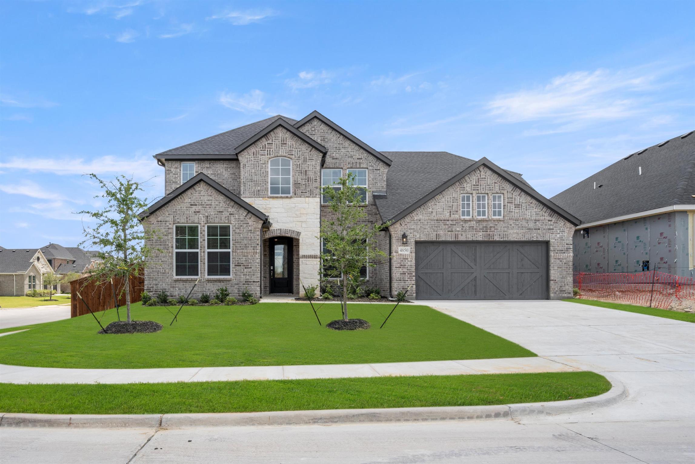 A two-story brick and stone house with a garage, surrounded by a well-manicured lawn and landscaping, set against a clear blue sky with scattered clouds.