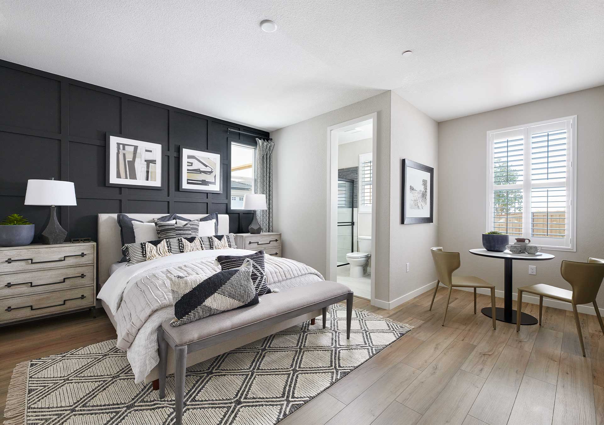 A modern and stylish bedroom with a gray accent wall, a large bed with patterned bedding, and a small dining area with a round table and chairs in the background.
