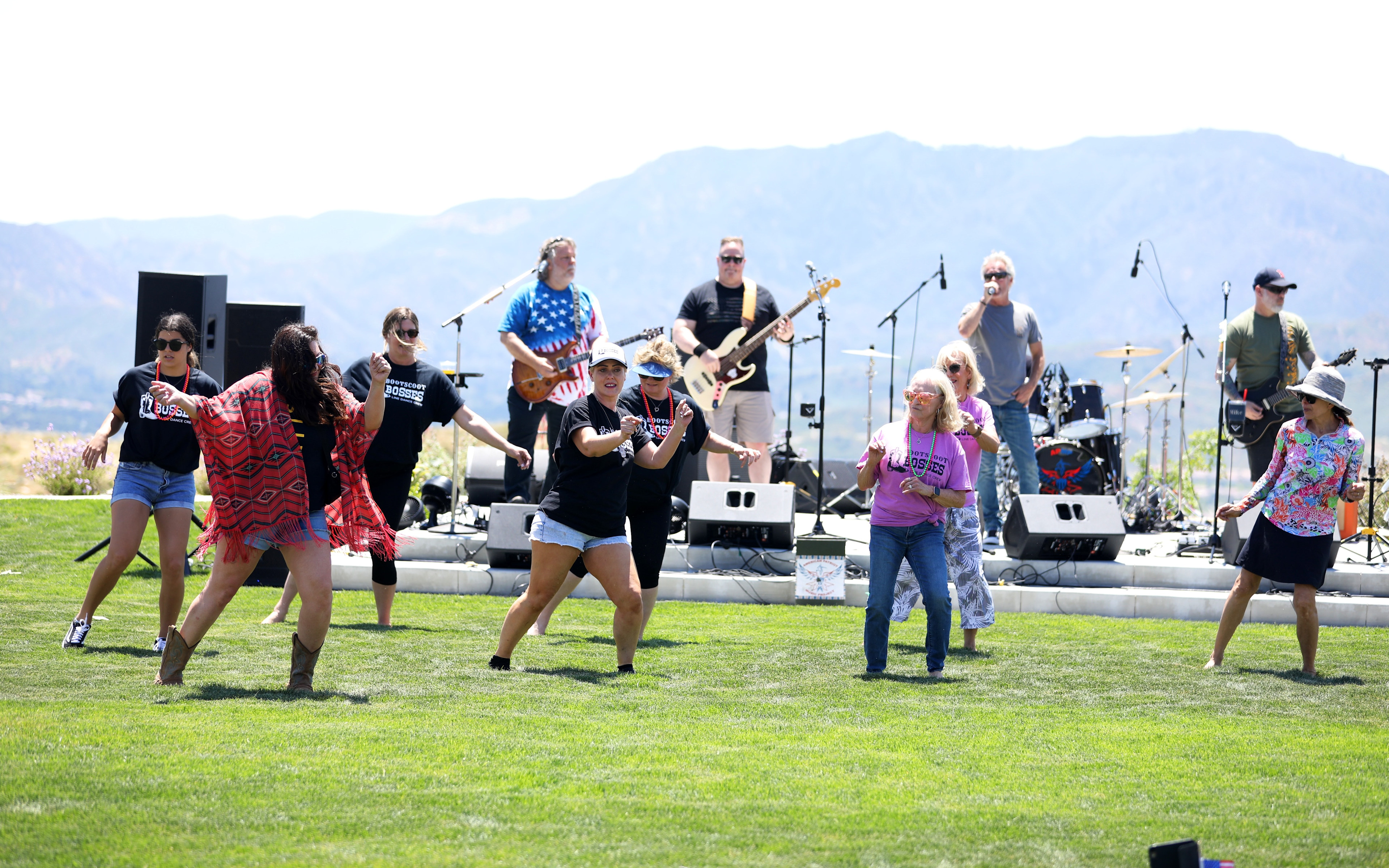 A group of people performing on a stage set up on a grassy field, with mountains visible in the background.
