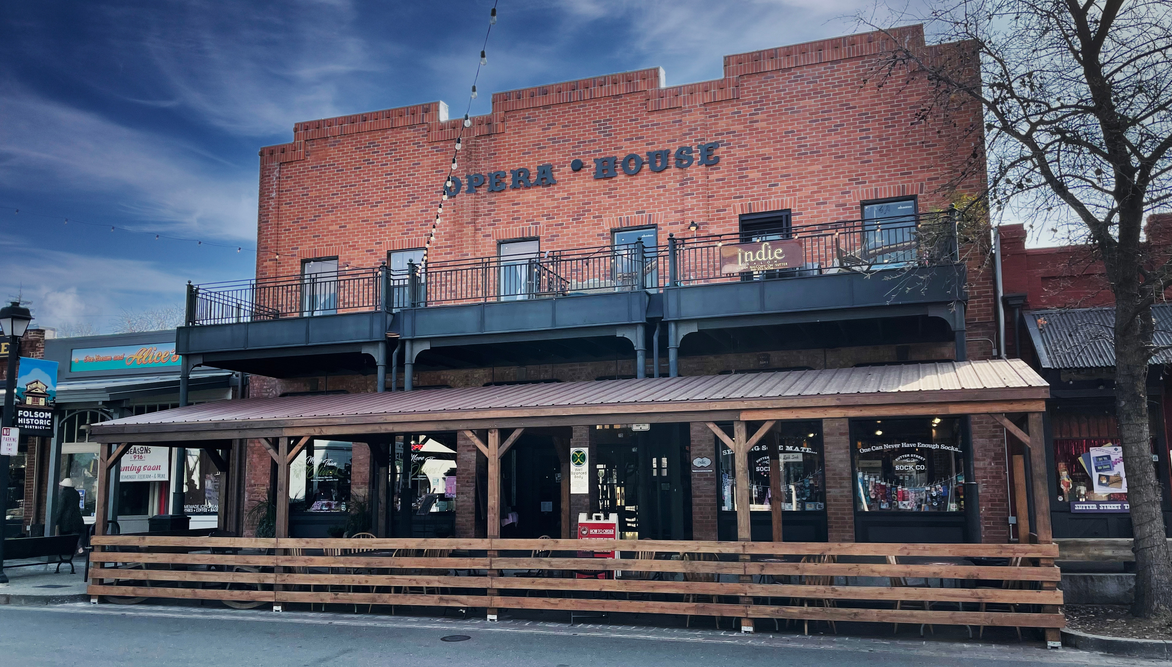 A brick building with the words "Opera House" prominently displayed, surrounded by a wooden deck and benches, set against a blue sky with some trees in the background.