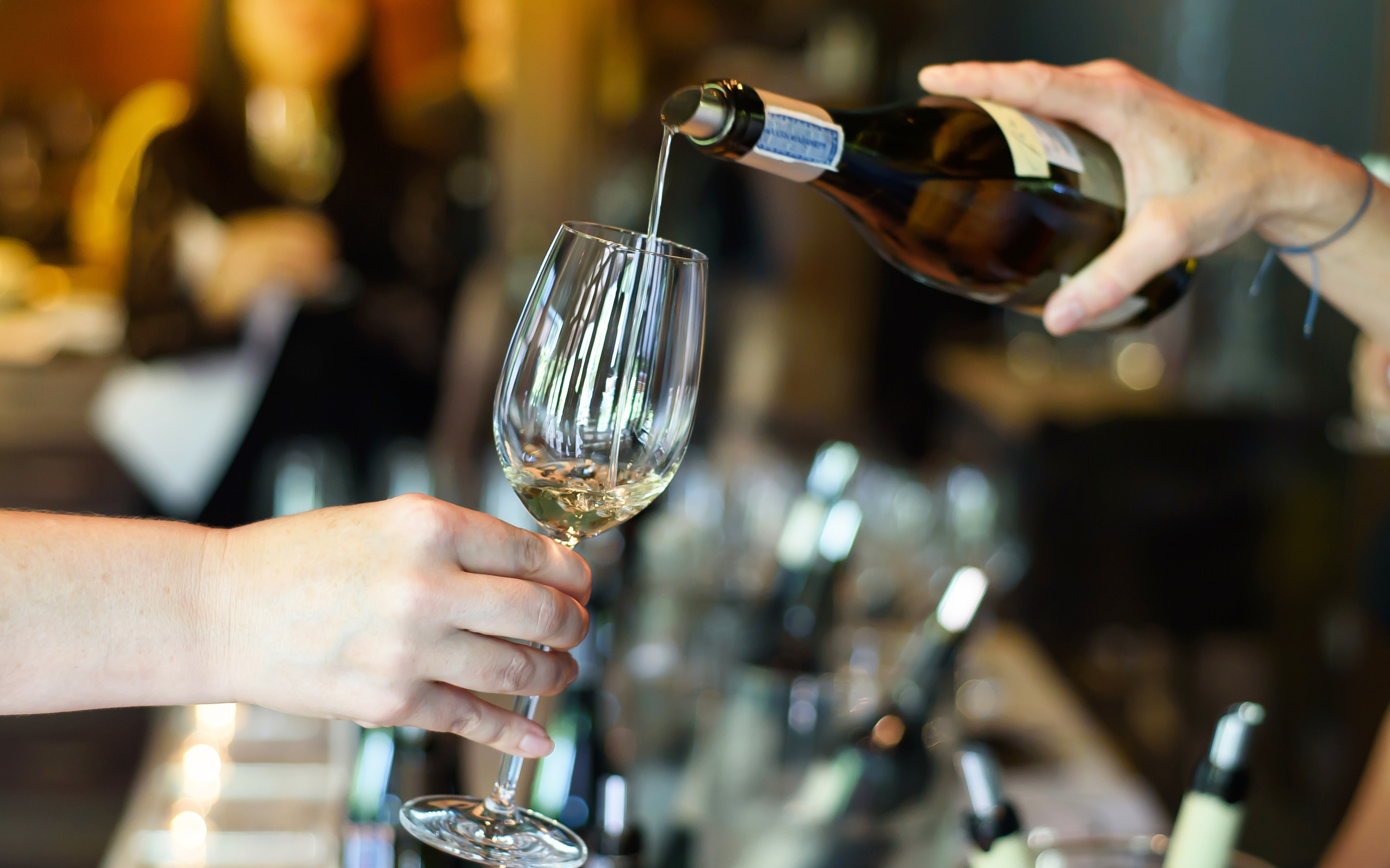A person's hands are pouring wine into a crystal wine glass against a backdrop of wine bottles and glasses.