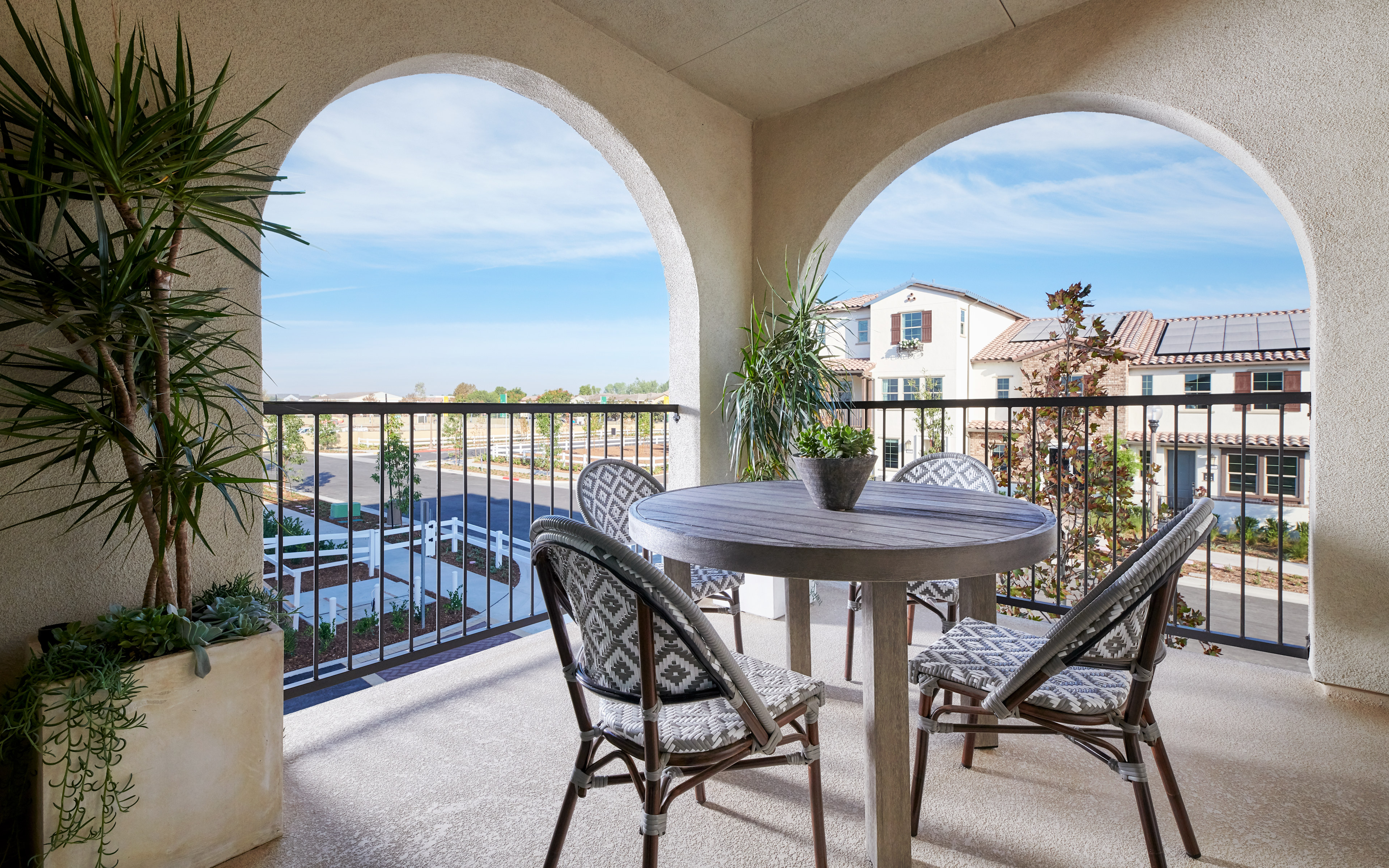 A cozy balcony with arched windows overlooking a scenic outdoor area, featuring a round table and chairs surrounded by potted plants.