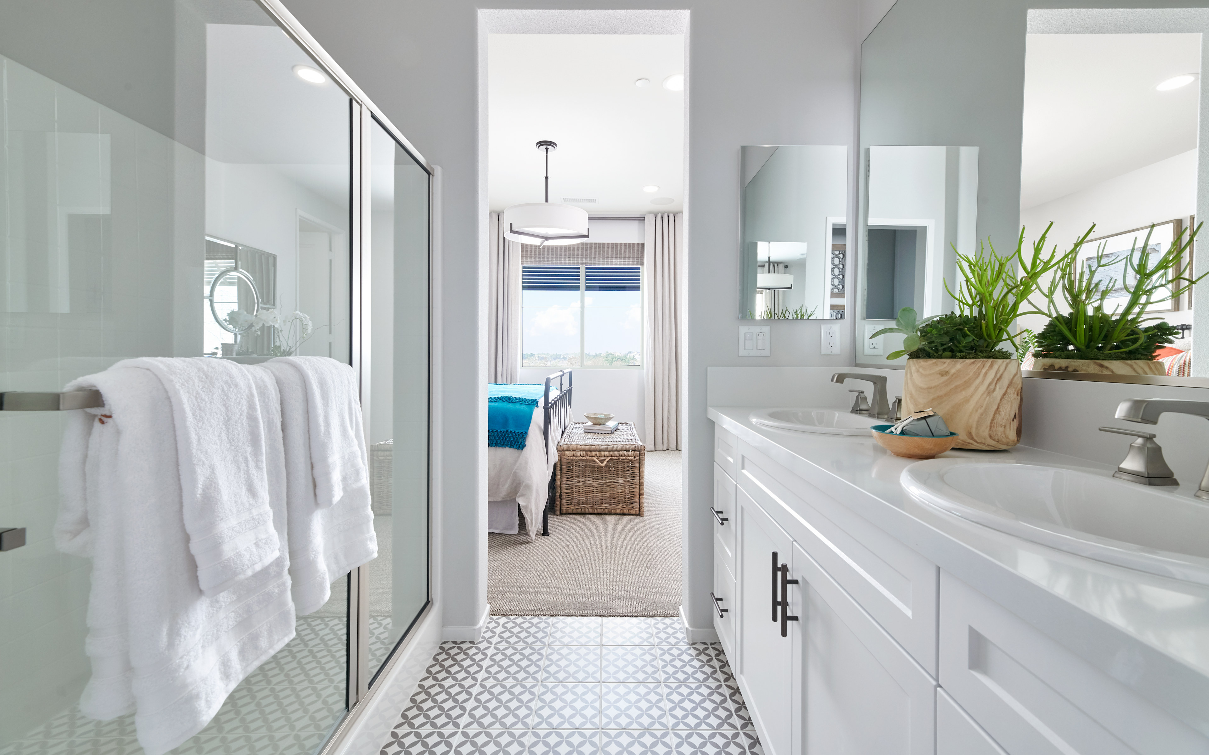 A bright and airy bathroom with white cabinets, a large mirror, and a potted plant, leading to an open doorway in the background.