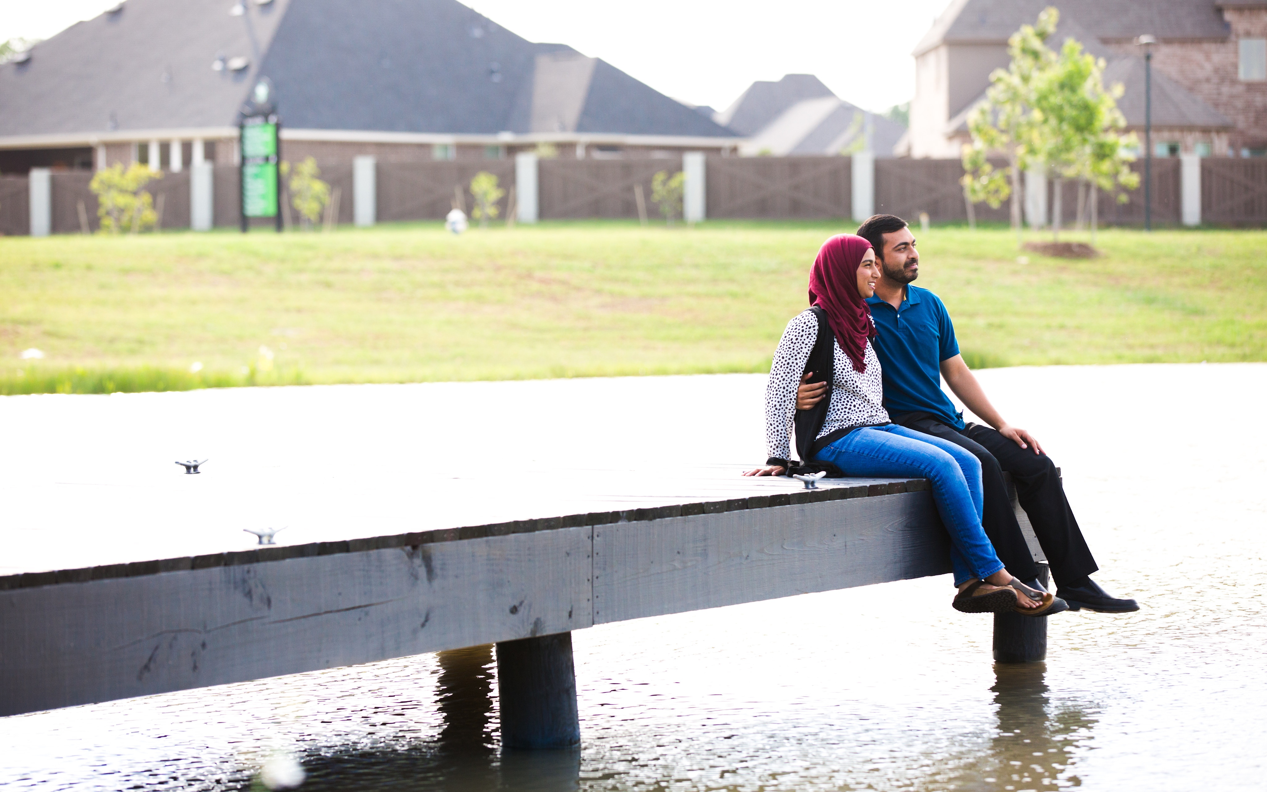 A couple sitting on a bench in a grassy area, with buildings and trees visible in the background.