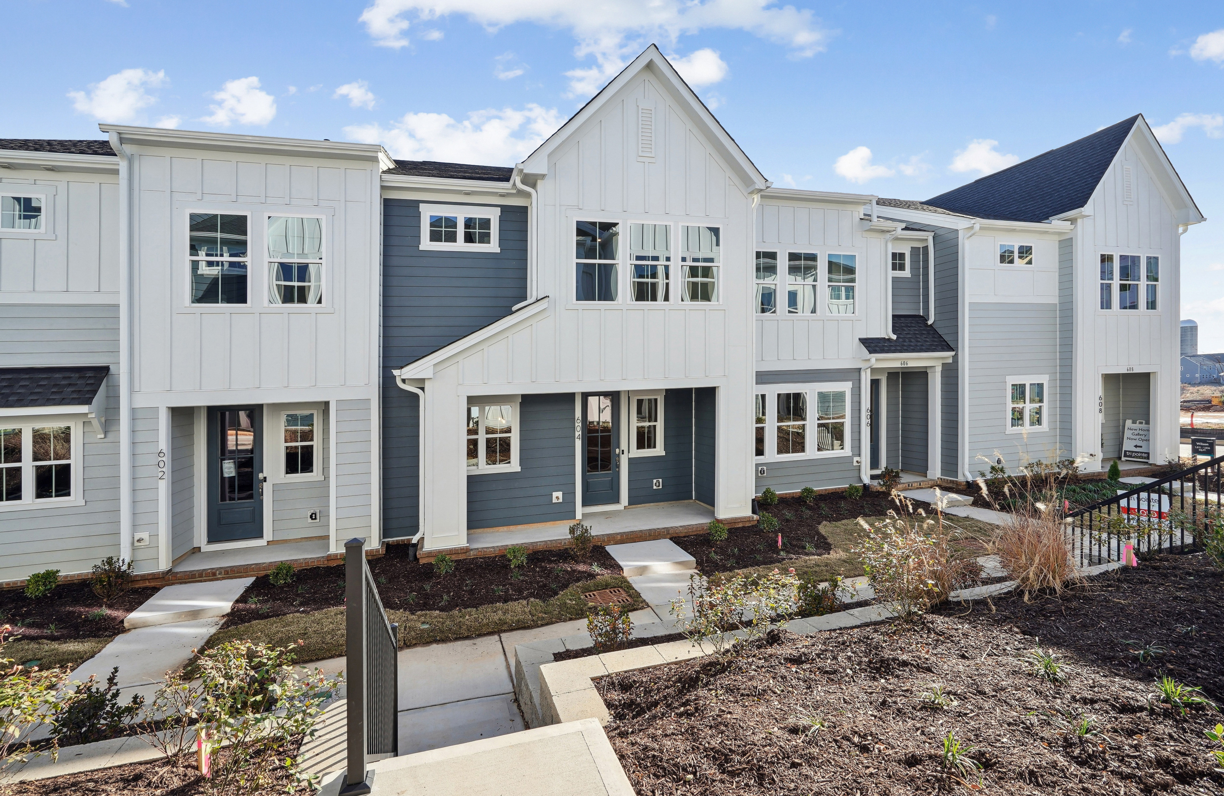 A row of modern, two-story townhouses with white siding, gray accents, and gabled roofs, set against a backdrop of a blue sky with scattered clouds.