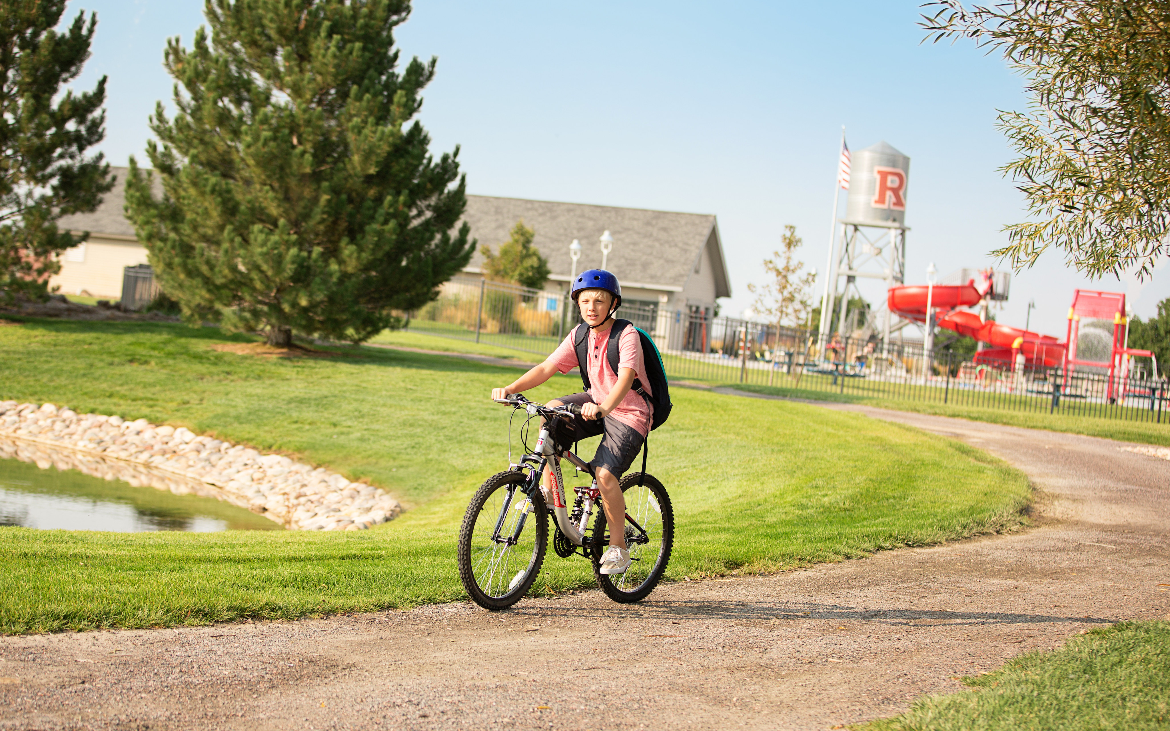A person wearing a blue hat and a pink jacket is riding a bicycle on a dirt path surrounded by lush greenery, with buildings and signage visible in the background.