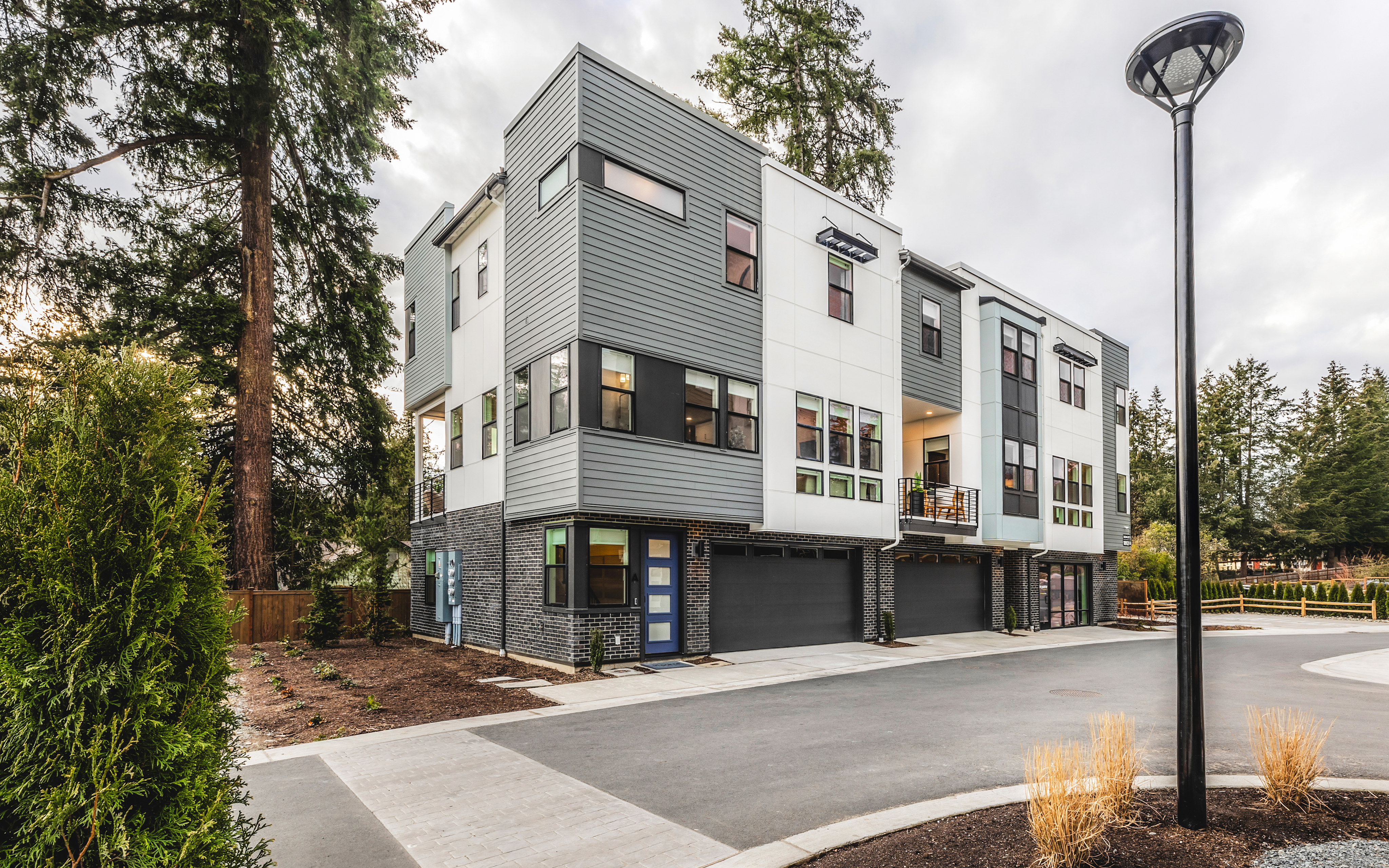 A modern, multi-story residential building with a gray exterior stands amidst a landscaped environment featuring trees, shrubs, and a paved walkway in the foreground.
