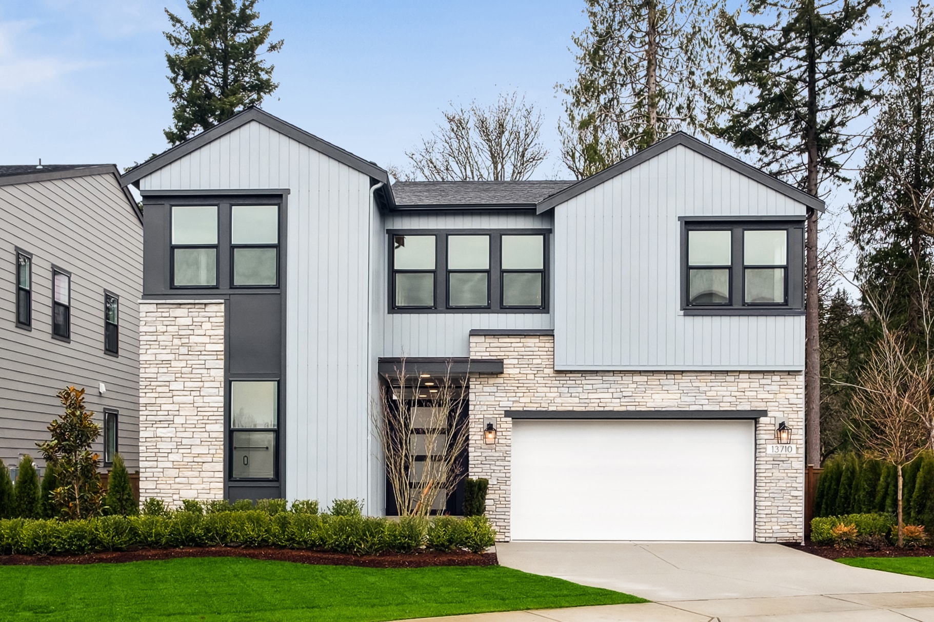 A modern, two-story house with a white exterior, black trim, and a garage door stands in a well-manicured yard surrounded by trees.
