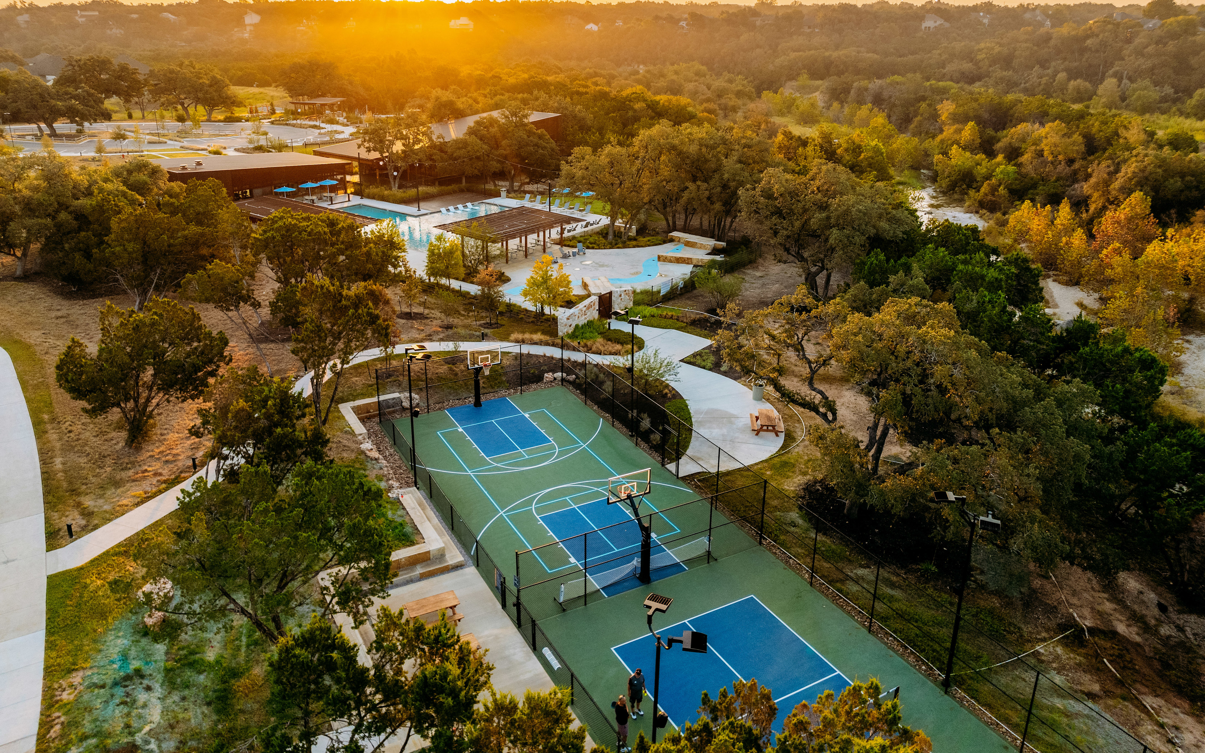 A scenic outdoor recreational area with tennis courts, swimming pools, and lush greenery, bathed in the warm glow of the setting sun.