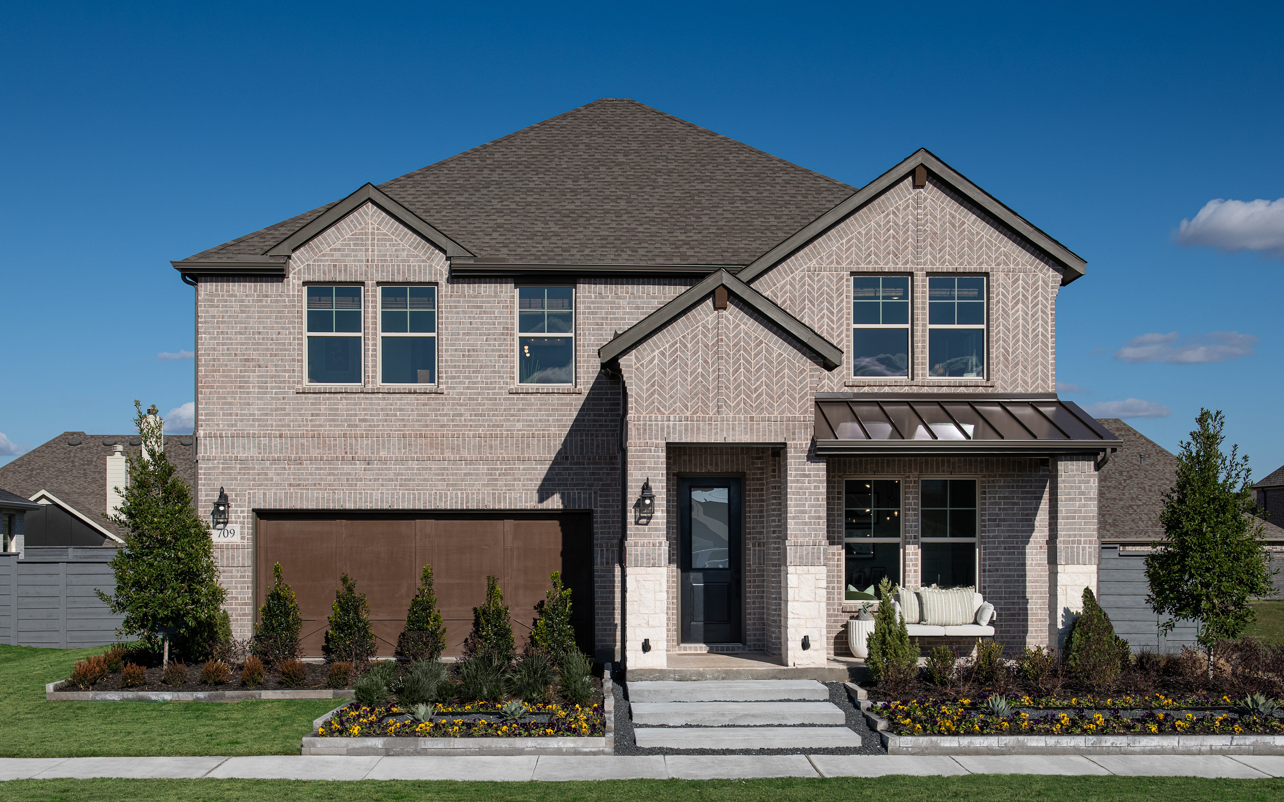 A two-story brick house with a gabled roof, surrounded by a well-manicured lawn and landscaping, stands against a clear blue sky.