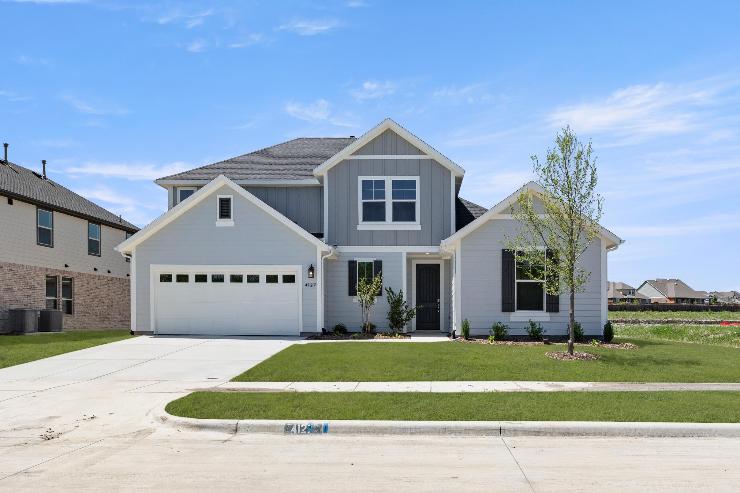 A two-story gray house with a garage, surrounded by a well-manicured lawn and trees in the background, stands in a residential neighborhood.