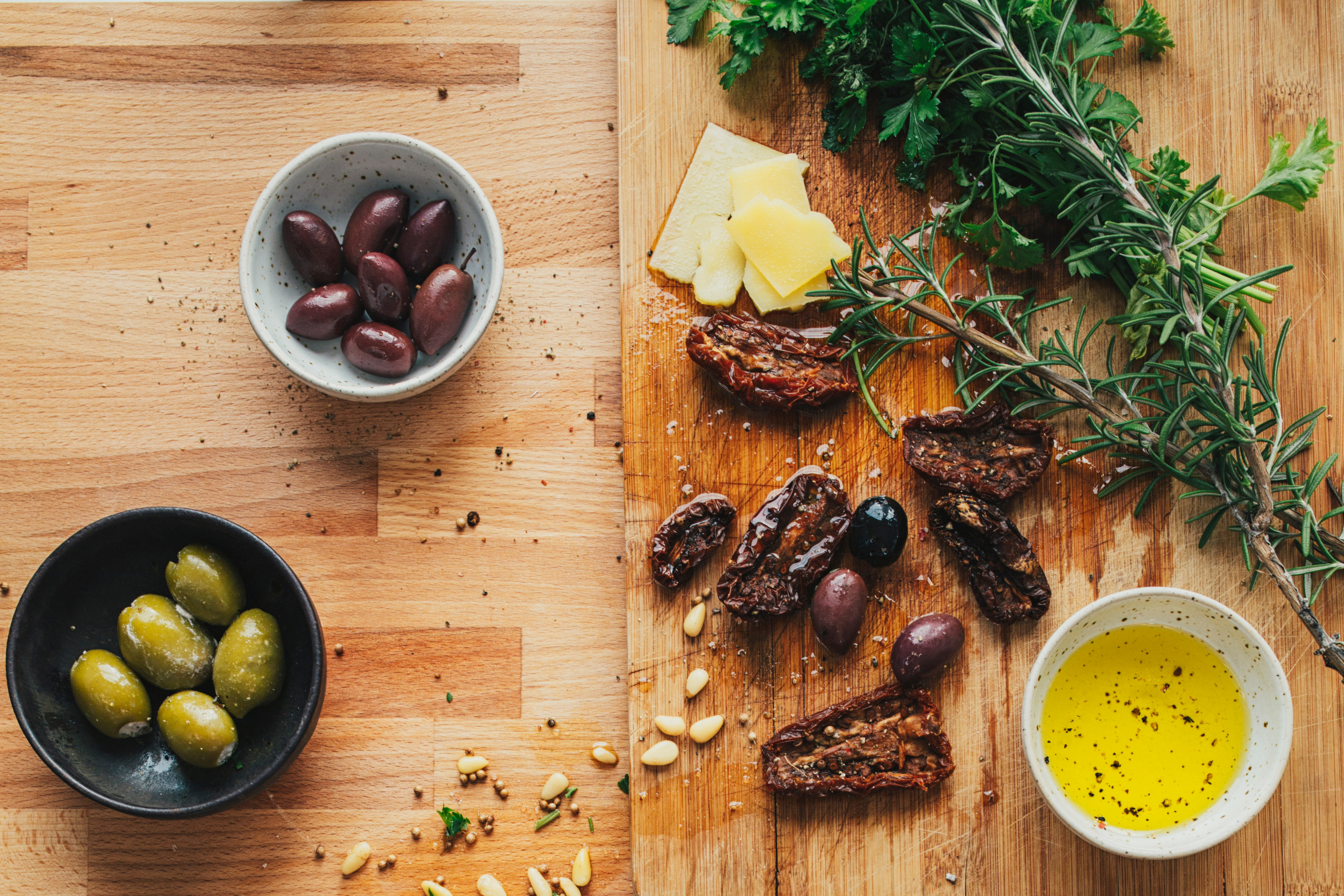 A wooden cutting board with various Mediterranean ingredients, including olives, dried fruits, herbs, and a person's hands preparing the food.