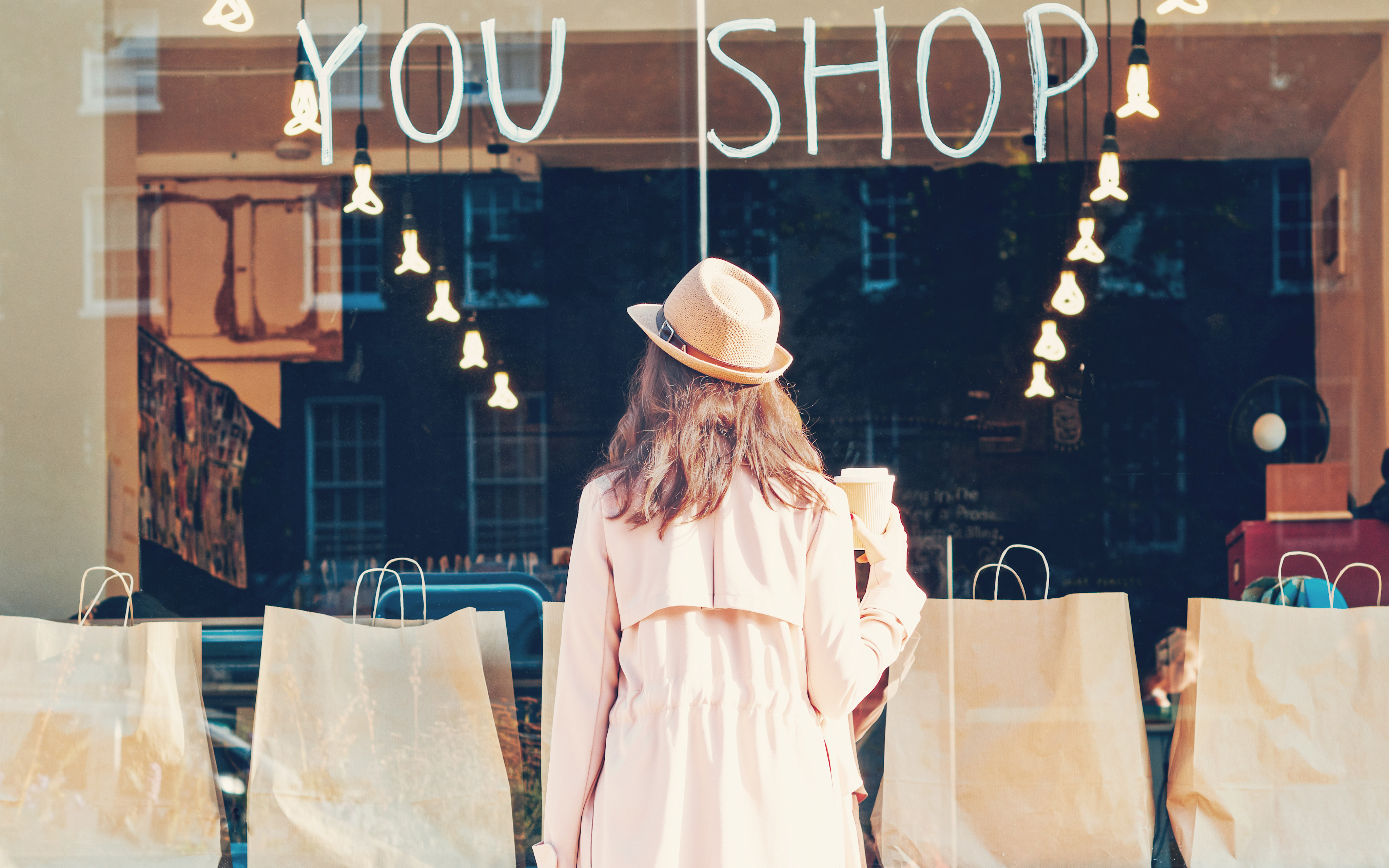 A woman in a light-colored coat and hat stands in front of a storefront with the words "YOU SHOP" displayed on the window, surrounded by shopping bags and a warm, inviting atmosphere.