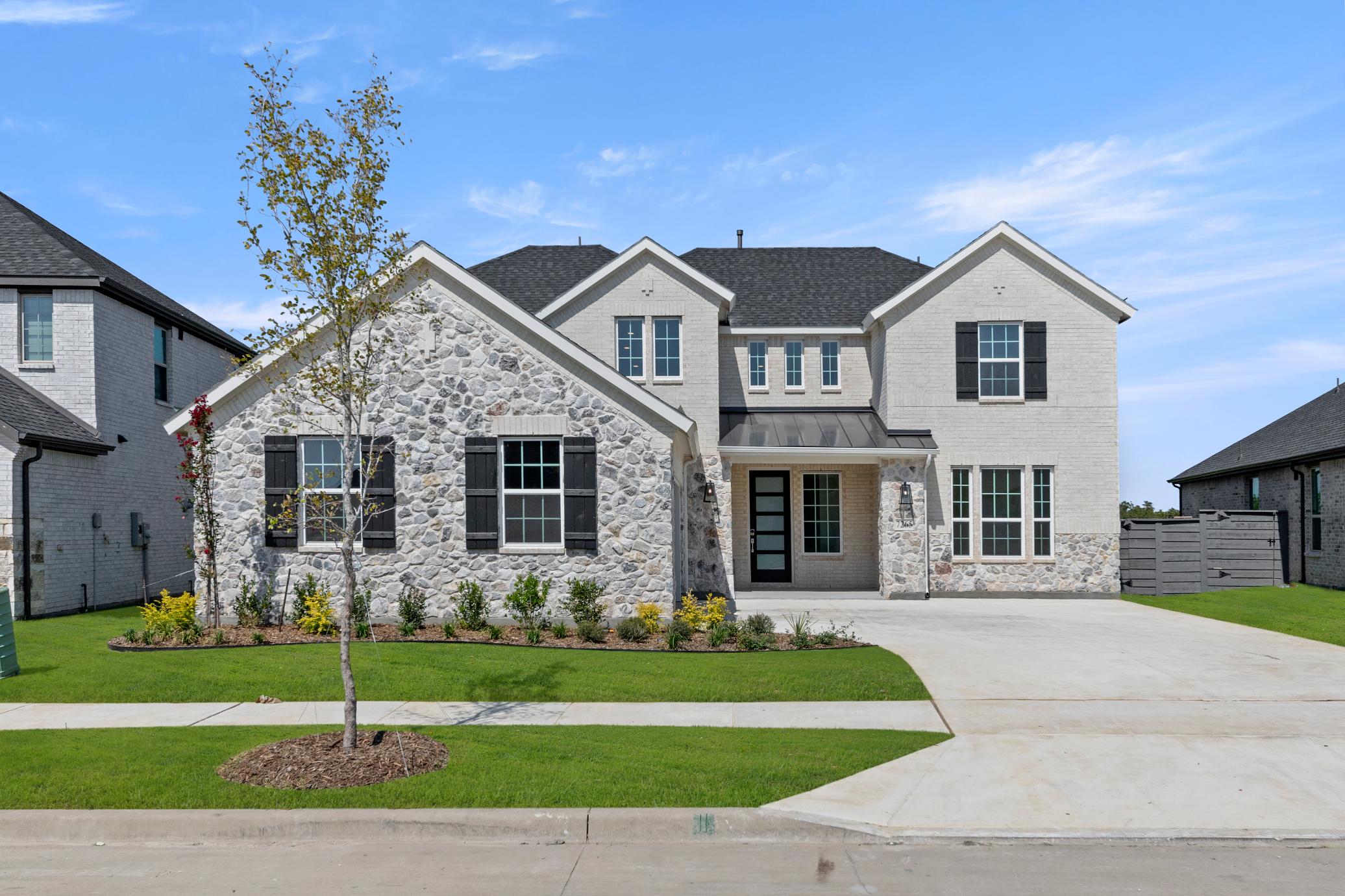 A large, two-story stone and wood-framed house with a well-manicured lawn and landscaping in the foreground, set against a clear blue sky with scattered clouds.