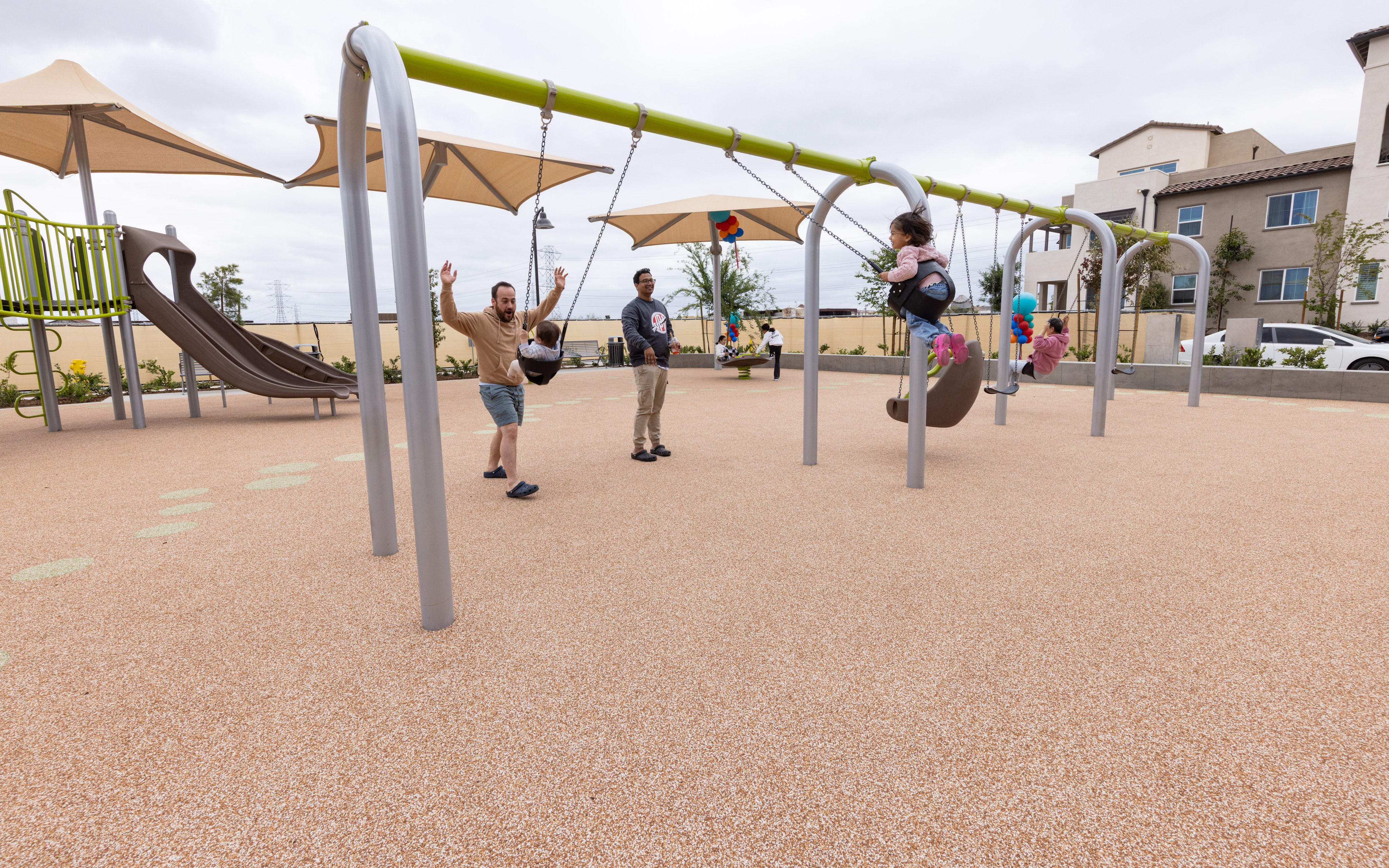 A modern outdoor playground with various colorful equipment and structures, surrounded by residential buildings and shaded areas.