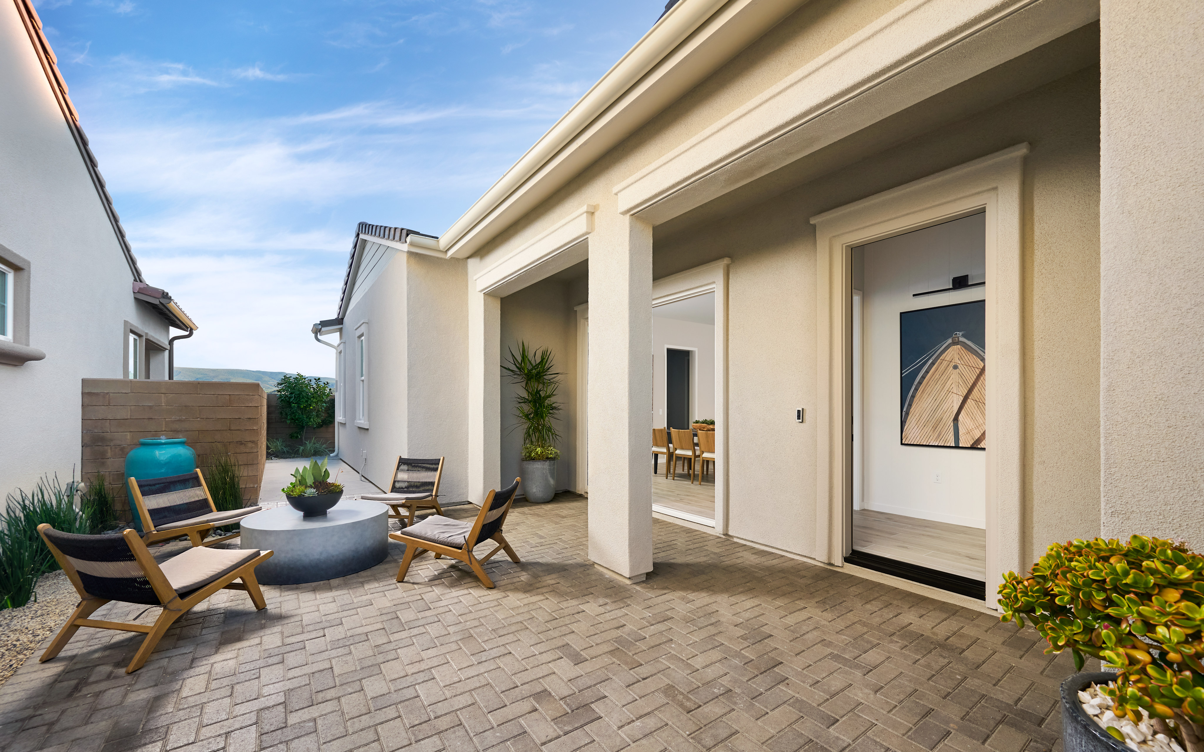 A cozy outdoor patio with wooden furniture, potted plants, and a brick-paved floor, surrounded by a modern, beige-colored house with large windows and an inviting entryway.