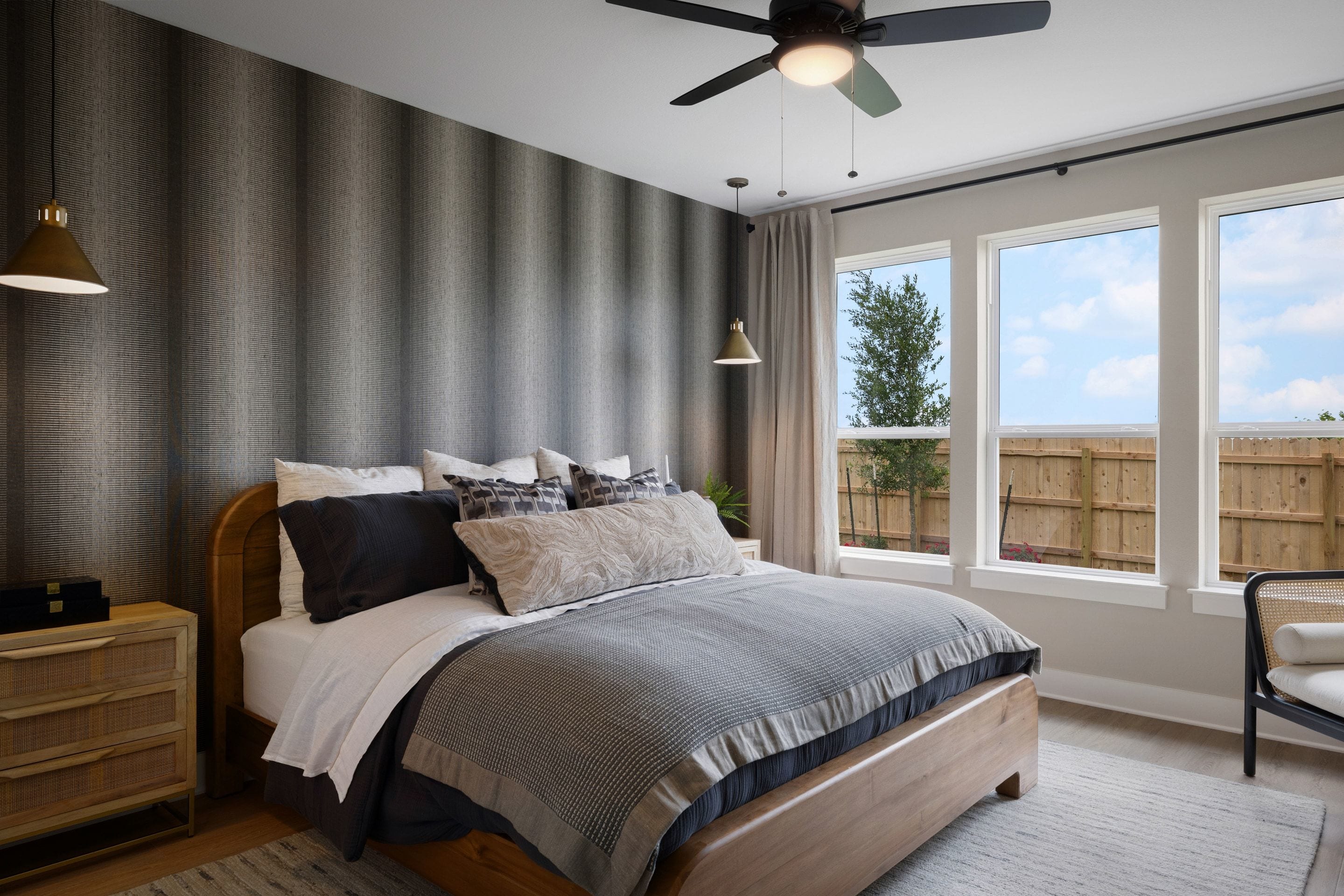 A modern bedroom with a plush bed in the foreground and large windows revealing a backyard with a fence and trees in the background.