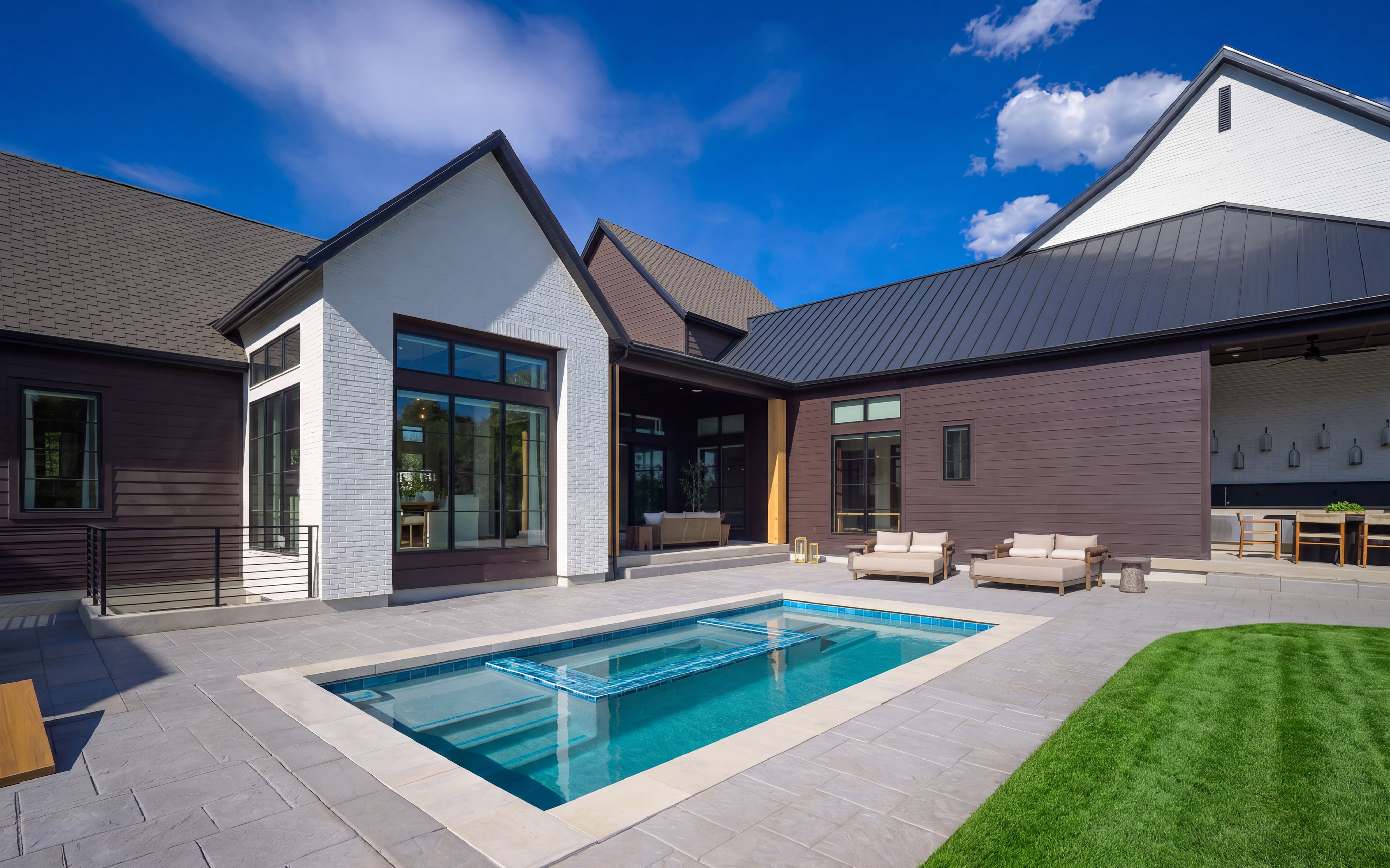 A rectangular swimming pool with a tiled insert sits on a paved patio in the foreground, while a modern house with dark siding and white accents, along with a manicured lawn, forms the background under a bright blue sky.