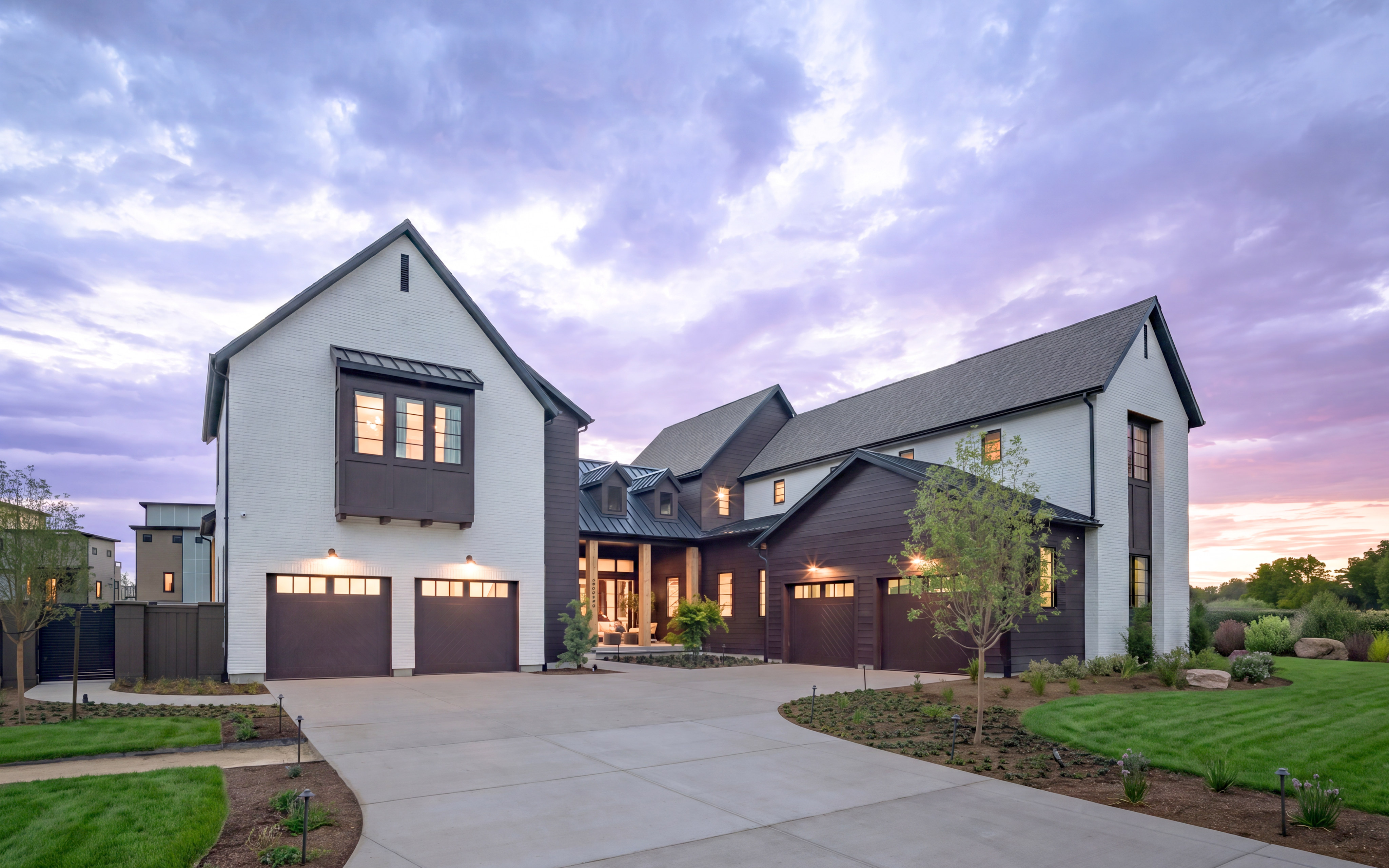A modern white and dark wood house with a wide driveway in the foreground sits under a dramatic purple and pink sky.