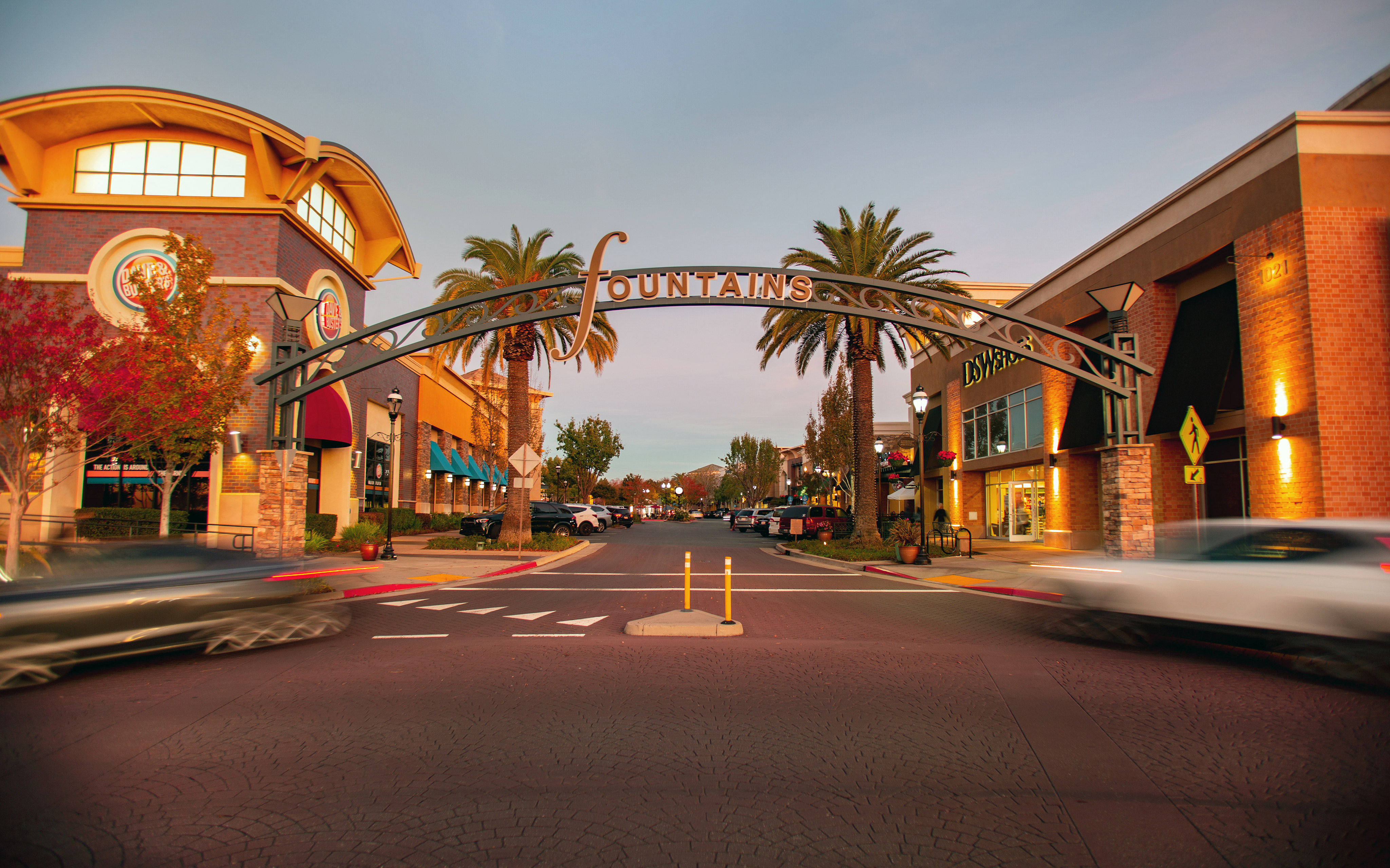 Blurred cars speed across a paved street in the foreground, leading toward a decorative "Fountains" archway flanked by palm trees and illuminated storefronts under a twilight sky.