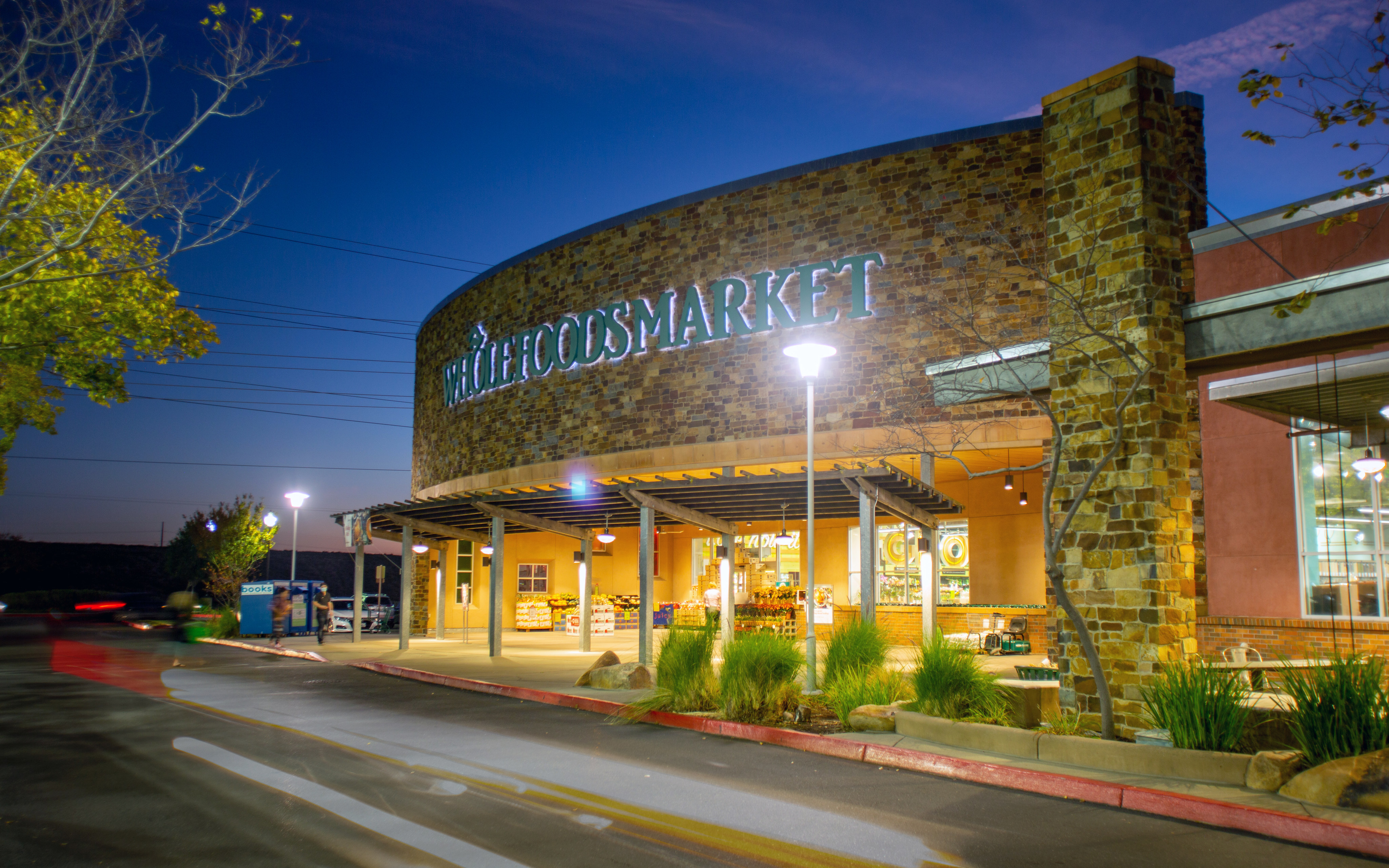 A stone-walled Whole Foods Market building stands under a deep blue twilight sky, with a paved road and small green shrubs in the foreground.