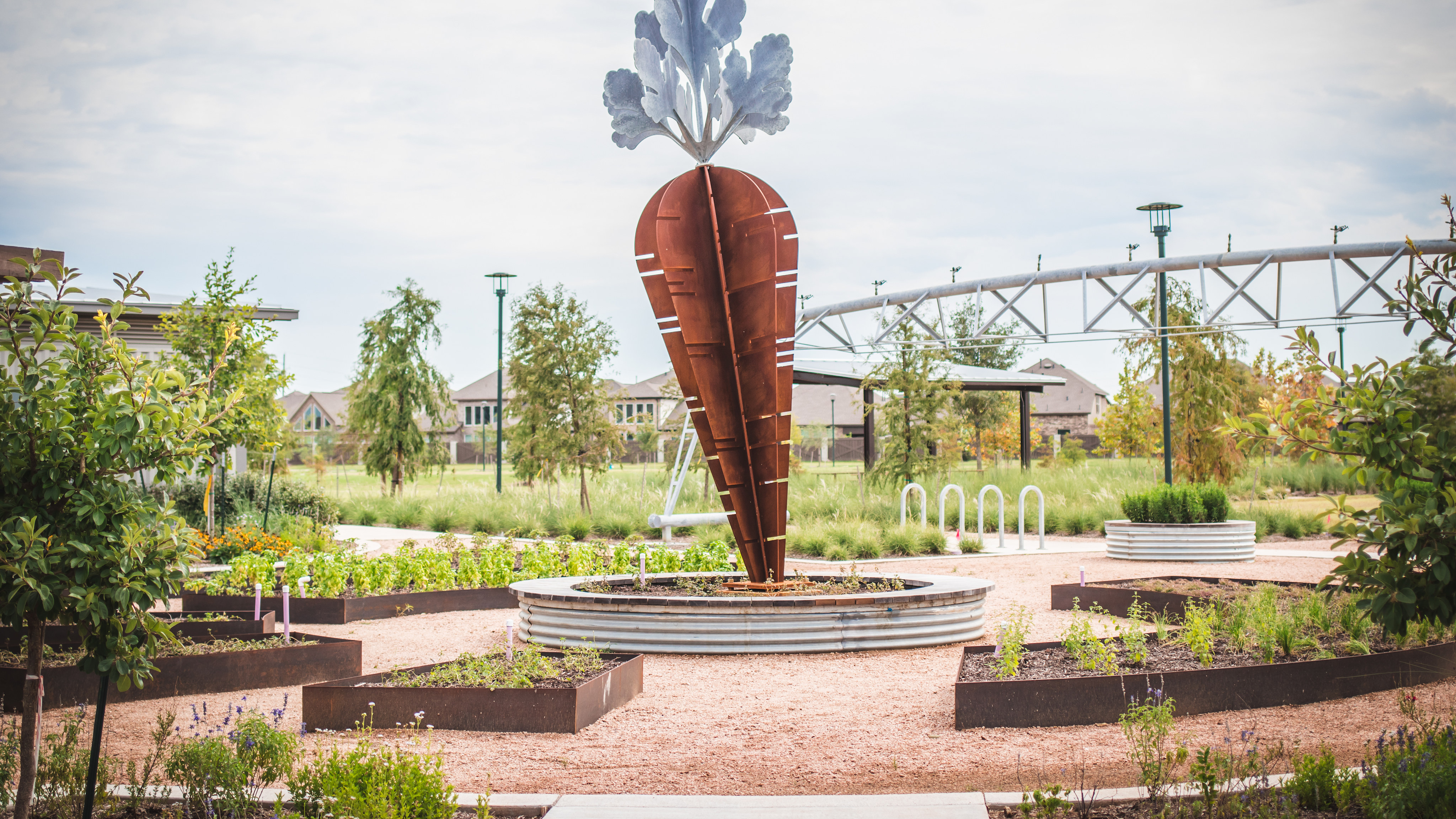 A large, red carrot-shaped sculpture stands prominently in a well-manicured garden, surrounded by lush greenery and architectural structures in the background.