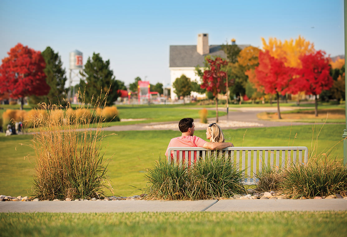 A peaceful park scene with a person sitting on a bench surrounded by vibrant autumn foliage and a grassy lawn, with buildings visible in the background.
