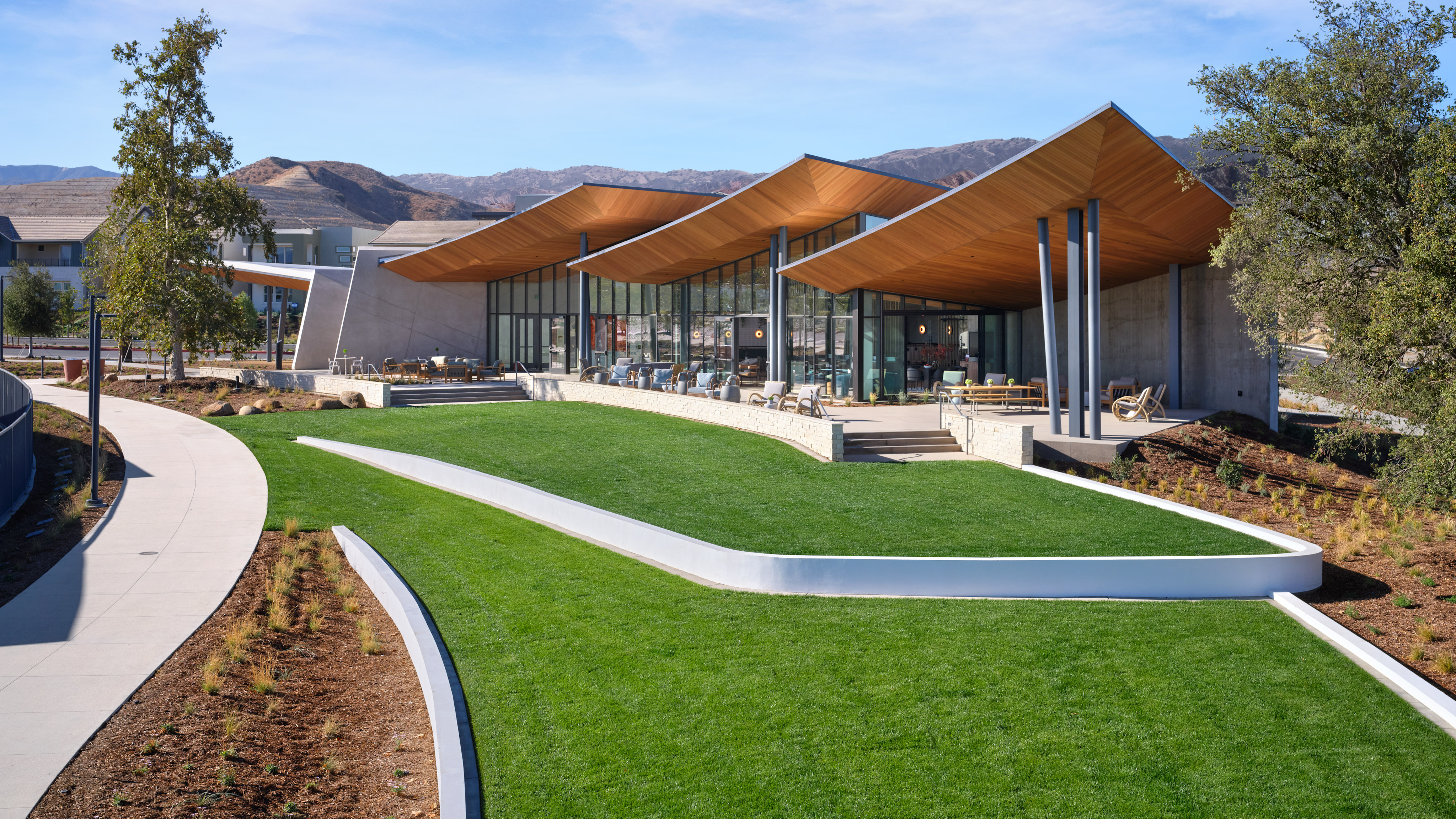 A modern, architecturally-designed building with a curved, orange-colored roof stands in the foreground, surrounded by a well-manicured lawn and landscaping. In the background, a mountainous landscape can be seen under a clear, blue sky.