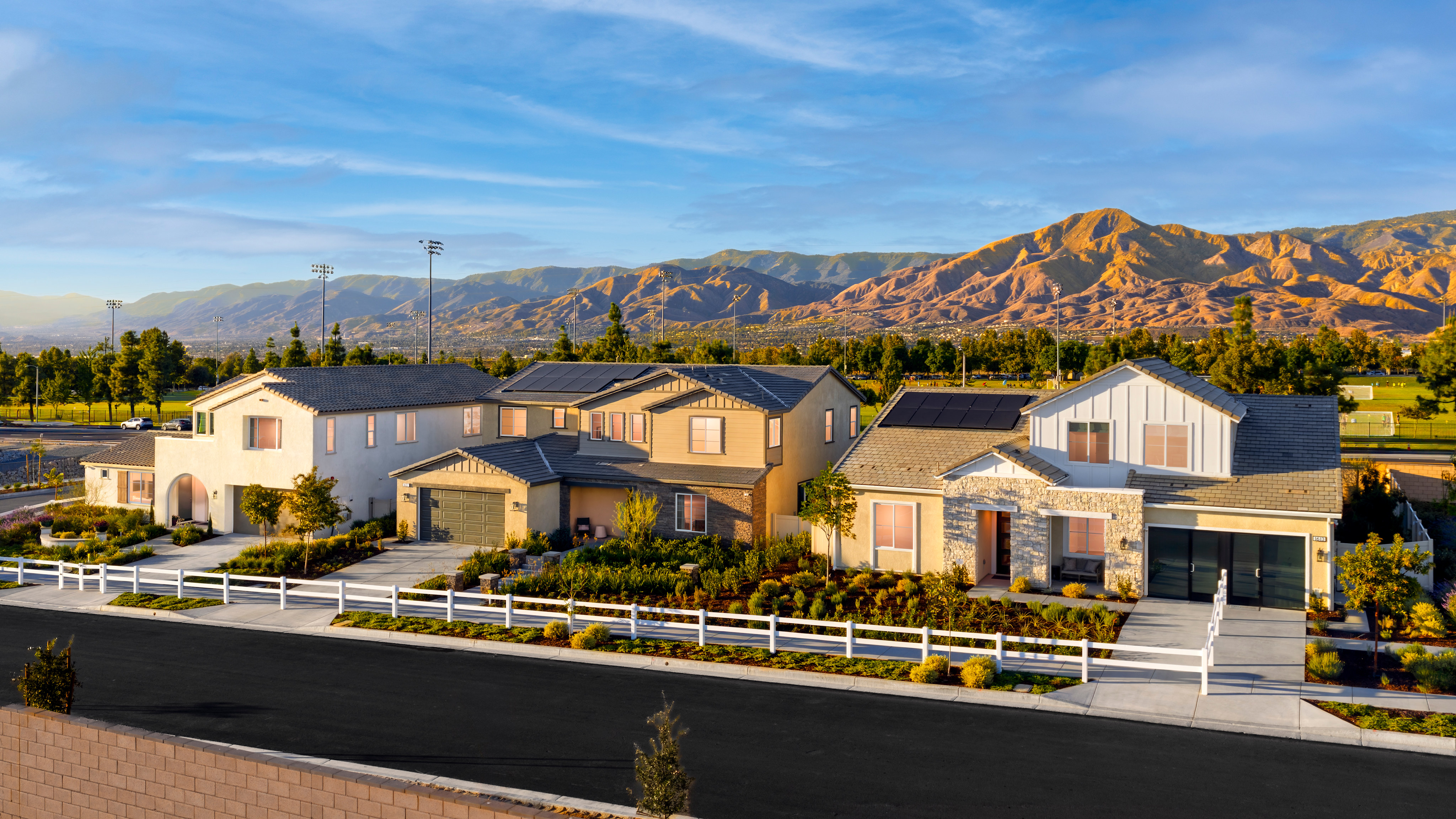 A residential neighborhood with modern houses nestled against a backdrop of rugged, sun-drenched mountains under a vibrant blue sky with wispy clouds.