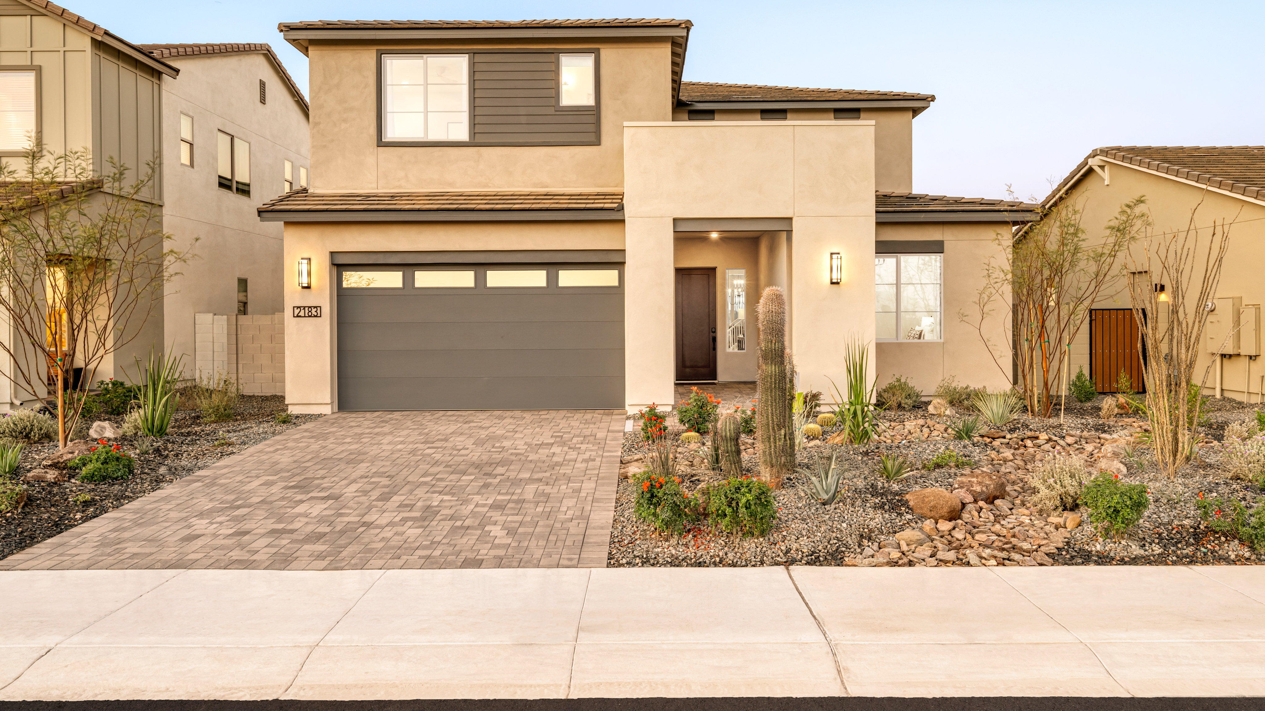 A two-story modern house with a garage, surrounded by a landscaped yard featuring various plants and a paved driveway.
